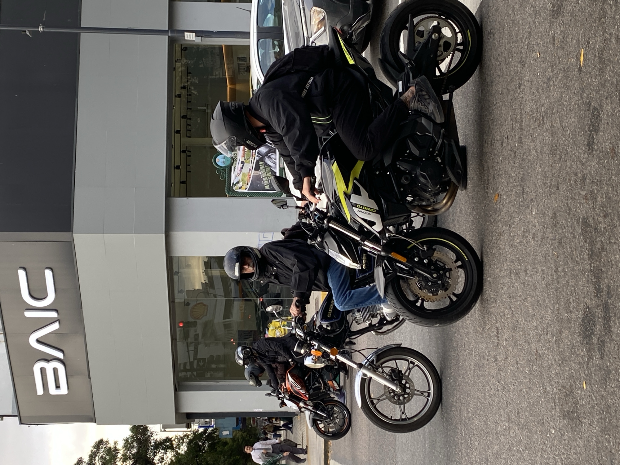 The image shows a row of motorcycles parked on a street in Buenos Aires, Argentina. Three motorcyclists wearing helmets are sitting on their bikes. The sign for the BAIC automotive company is visible on a building in the background. Pedestrians are walking on the sidewalk. The lighting suggests it is daytime.