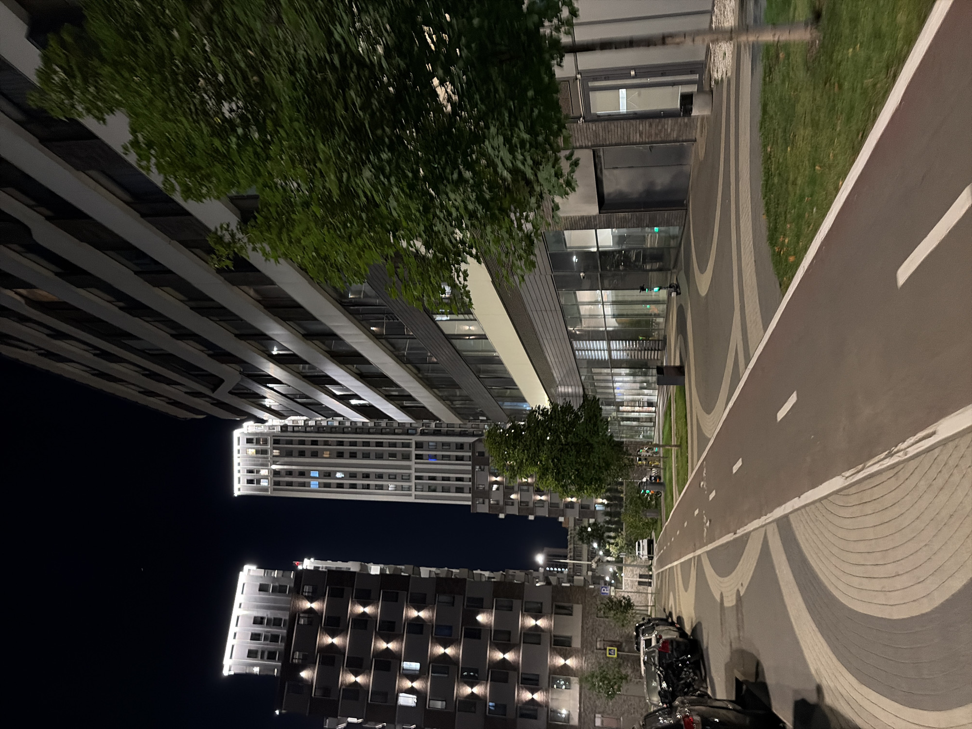 The image depicts a night scene in a modern residential complex, likely located in Moscow, Russia, given the architectural style and urban setting. In the foreground, a paved path with a red cycling lane is visible, bordered by a light-colored sidewalk. The path curves to the left, leading towards several multi-story apartment buildings. These buildings are contemporary in design, with dark facades and numerous illuminated windows. Some buildings feature decorative lighting at their upper levels.
To the left of the path, several cars are parked along the street. The ground paving in this area is a patterned mosaic of gray and light-colored stones. To the right of the path, there is a strip of green grass and a few trees. Beyond the grass and trees, the ground floor of another building features large glass windows, suggesting commercial or residential units. A few scooters are parked near the building entrance.
In the background, more apartment buildings of varying heights and designs are visible, all illuminated by internal lighting. The sky above is dark, indicating nighttime, with a faint crescent moon visible. The overall impression is of a well-maintained, modern urban environment.