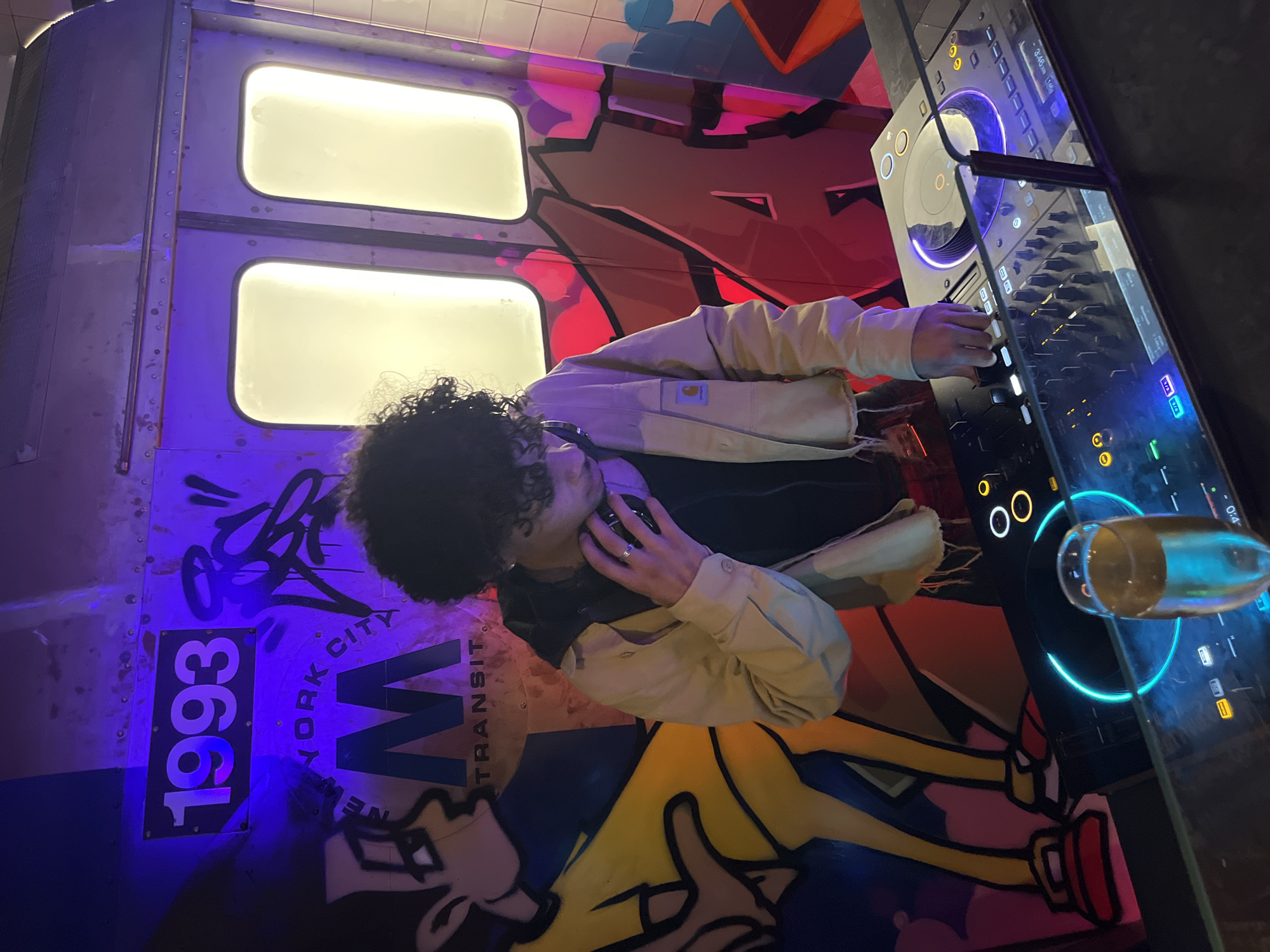 The image captures a DJ mixing music at a venue in Barcelona, Spain. The DJ, positioned on the left side of the frame, operates a sophisticated mixing console. A glass containing liquid, likely a beverage, rests on the console's surface.
The setting is characterized by an artistic backdrop resembling a New York City subway car exterior. The design includes graffiti elements and references the "New York City Transit" system, along with the year "1993". The backdrop's color palette incorporates blue, yellow, red, and grey tones.