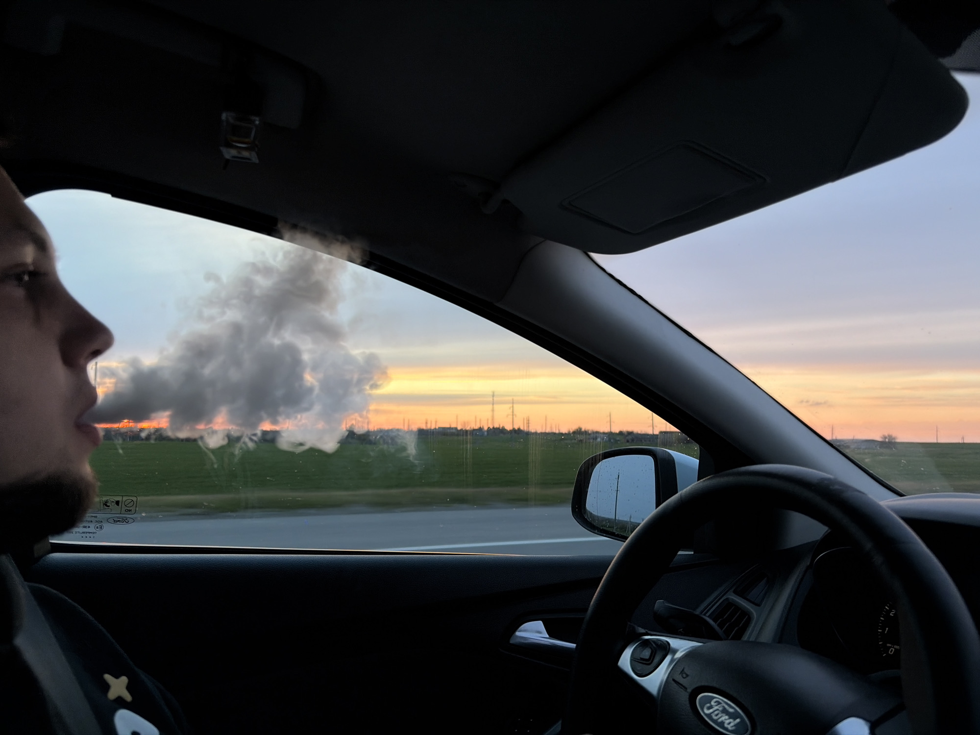The image captures a scene from inside a car during either sunset or sunrise in Alexandrovka, Russia. On the left, a person is seen in profile, looking out the window. A distinct, large cloud of smoke or vapor is exhaling from their mouth and dispersing outside the car.
The car's interior features a dark dashboard, a steering wheel with a visible "Ford" logo, and a rearview mirror reflecting the passing scenery and showing small droplets. A sun visor is folded up towards the ceiling.
Outside the car, the sky is painted with warm hues of orange, pink, and light blue, characteristic of dawn or dusk. Green fields stretch towards the horizon, where multiple power lines or industrial towers are visible. A road runs alongside the fields.
Textual details include the "Ford" logo on the steering wheel. On the car window behind the person, Russian words are visible, appearing to be safety warnings like "БЕЗ РЕМНЯ" (without a seatbelt) and "НЕ ОТКРЫВАТЬ" (do not open). The overall scene depicts a tranquil moment of travel, with the act of exhaling smoke/vapor contrasted against the serene, colorful sky.