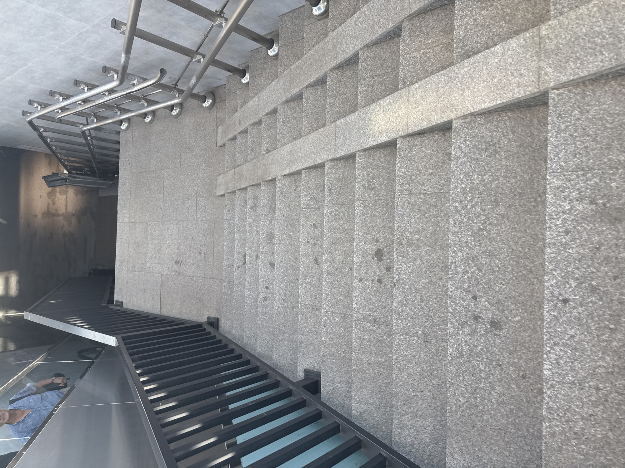 The image shows a downward view of a wide, outdoor staircase. The steps are made of speckled grey granite. On the left side of the staircase, there is a black metal railing with vertical bars. On the right side, there is a polished metal railing with horizontal bars, supported by circular bases. Two parallel ramps made of the same grey granite are present on the staircase, extending downwards. At the top of the staircase, on the left, an escalator is partially visible, with a man in a blue plaid shirt and dark pants walking down it. He is carrying a dark bag over his right shoulder. The environment appears to be a public transit area or an underpass in Bishkek City, Kyrgyzstan.