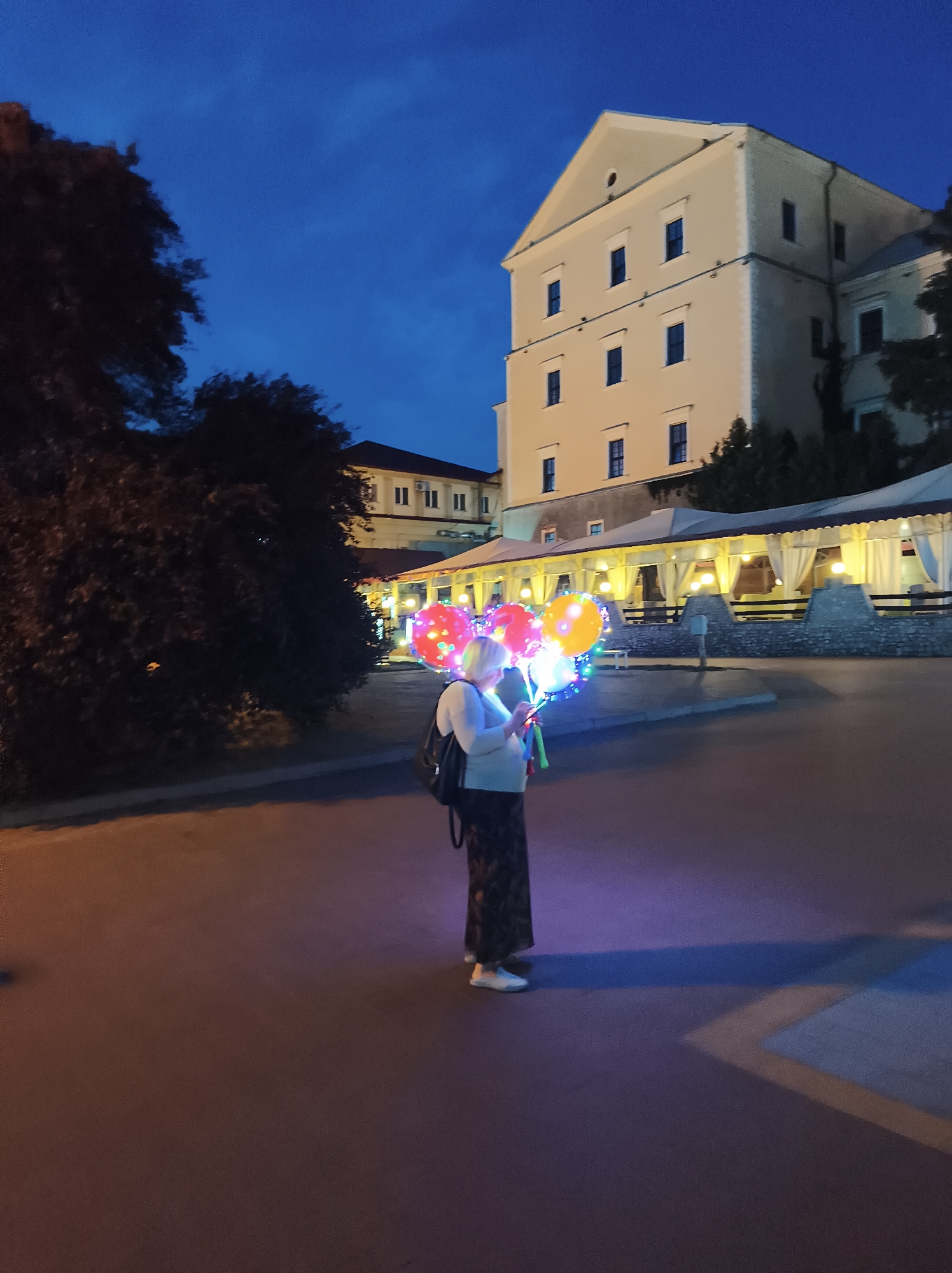 The image captures a twilight scene in the city of Ternopil, Ukraine, near the Ternopil Regional State Administration building. A woman stands on a paved pathway, holding a bunch of illuminated balloons. The balloons glow with various colors including red, yellow, blue, and green, with some featuring heart patterns. She is wearing a light-colored top, a dark patterned skirt, and white sneakers, and has a backpack slung over her shoulder. Behind her, the Ternopil Regional State Administration building is visible, characterized by its light-colored facade, numerous windows, and a prominent triangular roof. Adjacent to the building is an outdoor seating area with covered tents illuminated by warm light. The sky is a deep blue, indicating the progression from day to night. The overall atmosphere is festive and serene, with the illuminated balloons adding a touch of magic to the urban landscape.