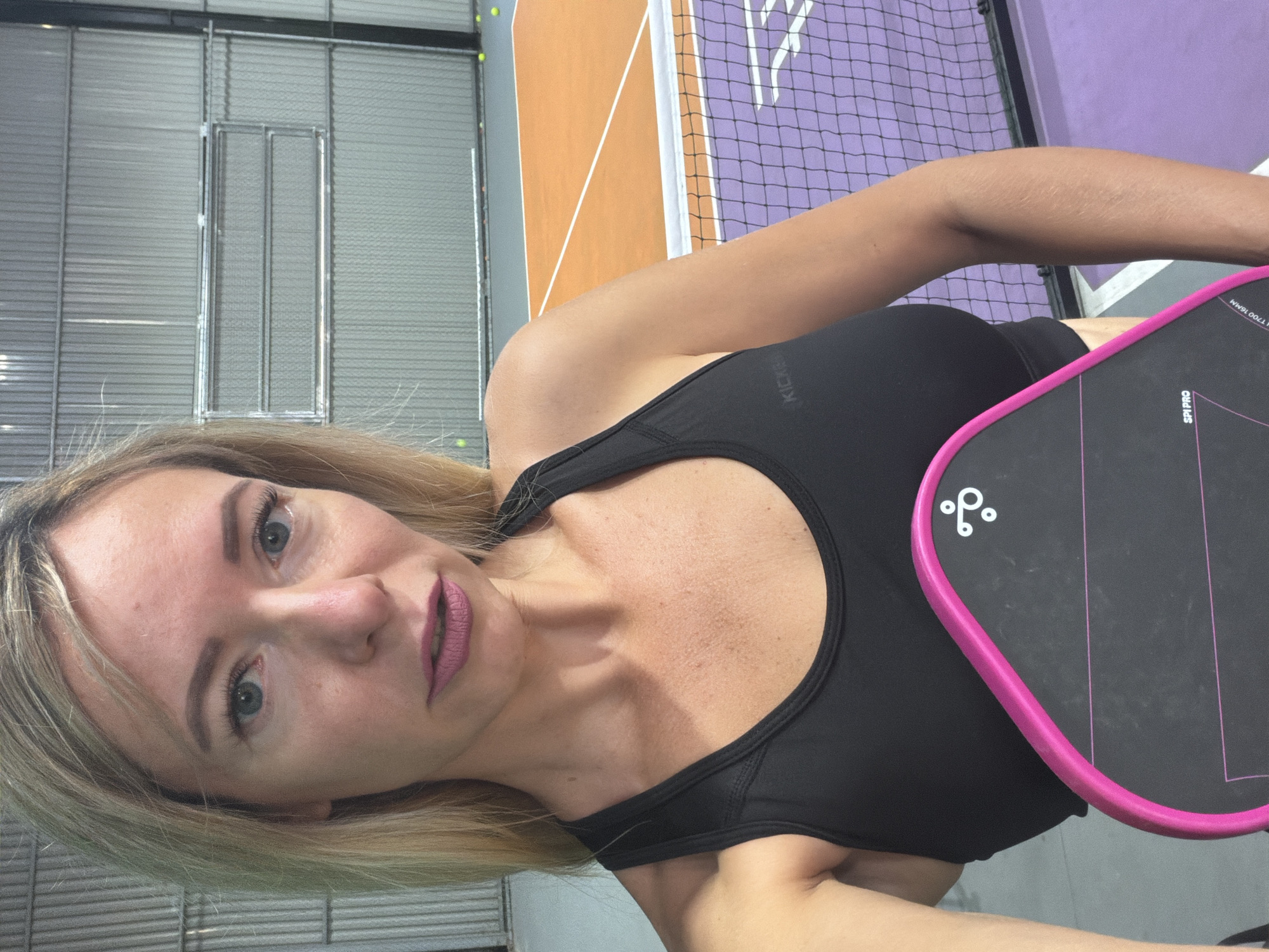 A blonde woman in a black sports bra stands indoors holding a pink and black paddle. She is facing the camera with a neutral expression, her hair is shoulder-length and blonde. In the background, there is a purple pickleball court with orange accents. The walls are corrugated metal. There are several yellow pickleballs visible on the court. The location is an indoor pickleball court.