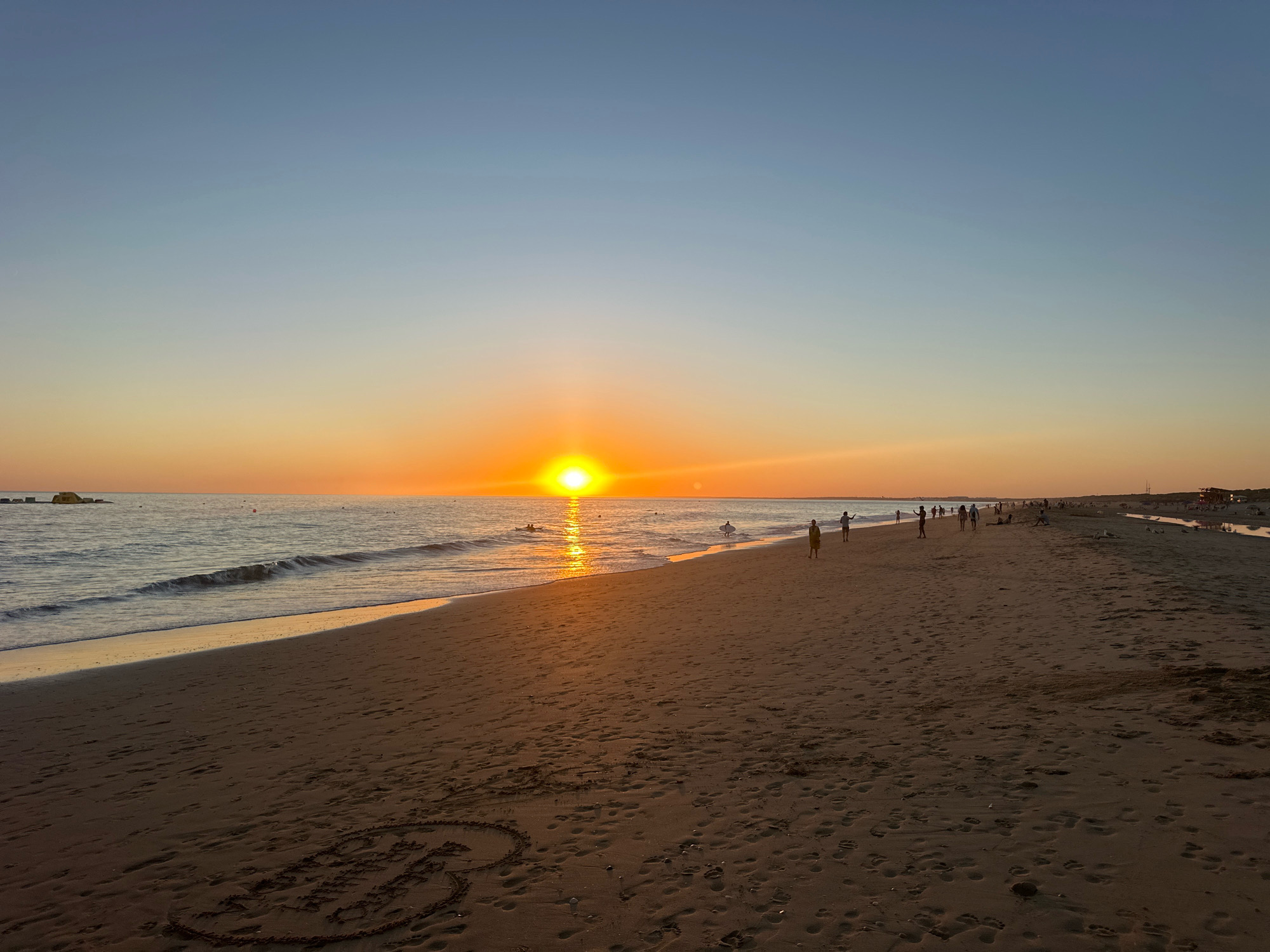 The image depicts a wide, sandy beach in Punta Umbría, Spain, during sunset. The sun is a bright, round orb low on the horizon, casting a warm orange glow across the sky and reflecting on the gentle waves of the sea. The water is a calm, shimmering blue, with small waves lapping at the shore. The beach itself is a warm brown color, with numerous footprints imprinted in the sand. In the foreground, a heart shape has been drawn in the sand.
Several people are scattered across the beach. Some are walking along the shoreline, while others are closer to the water. A few individuals are wading or standing in the shallow sea. Further down the beach, a small group appears to be gathered near some structures, possibly beach huts or facilities. In the distance, beyond the people, a channel of water is visible, possibly leading to or from the sea. The overall impression is a peaceful evening scene on a popular beach.