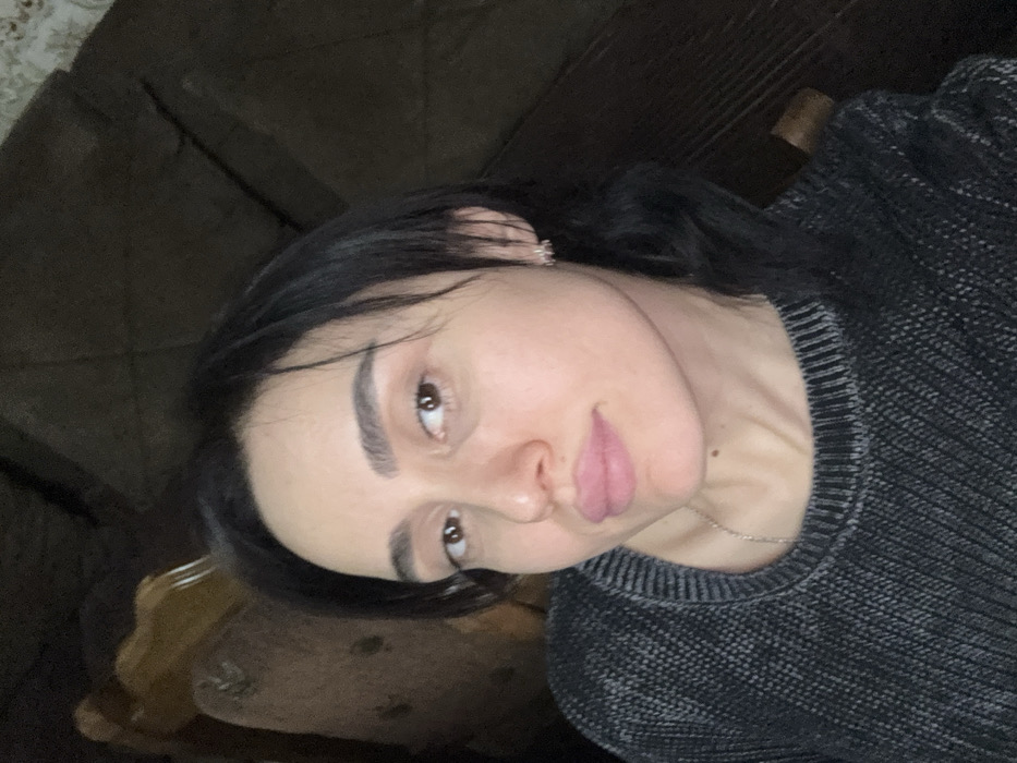 The scene depicts an adult female in an indoor setting within the city of Bolnisi, Georgia. The subject has dark hair, brown eyes, and fair skin, with light pink lipstick on her lips. She is wearing a dark grey, ribbed knit sweater, a thin silver-colored necklace, and a small, light-colored earring in her right ear. Her gaze is directed towards the camera. The background includes residential furniture: a wooden chair with an upholstered section is partially visible to the subject's left, and a dark upholstered piece of furniture is partially visible to her right.