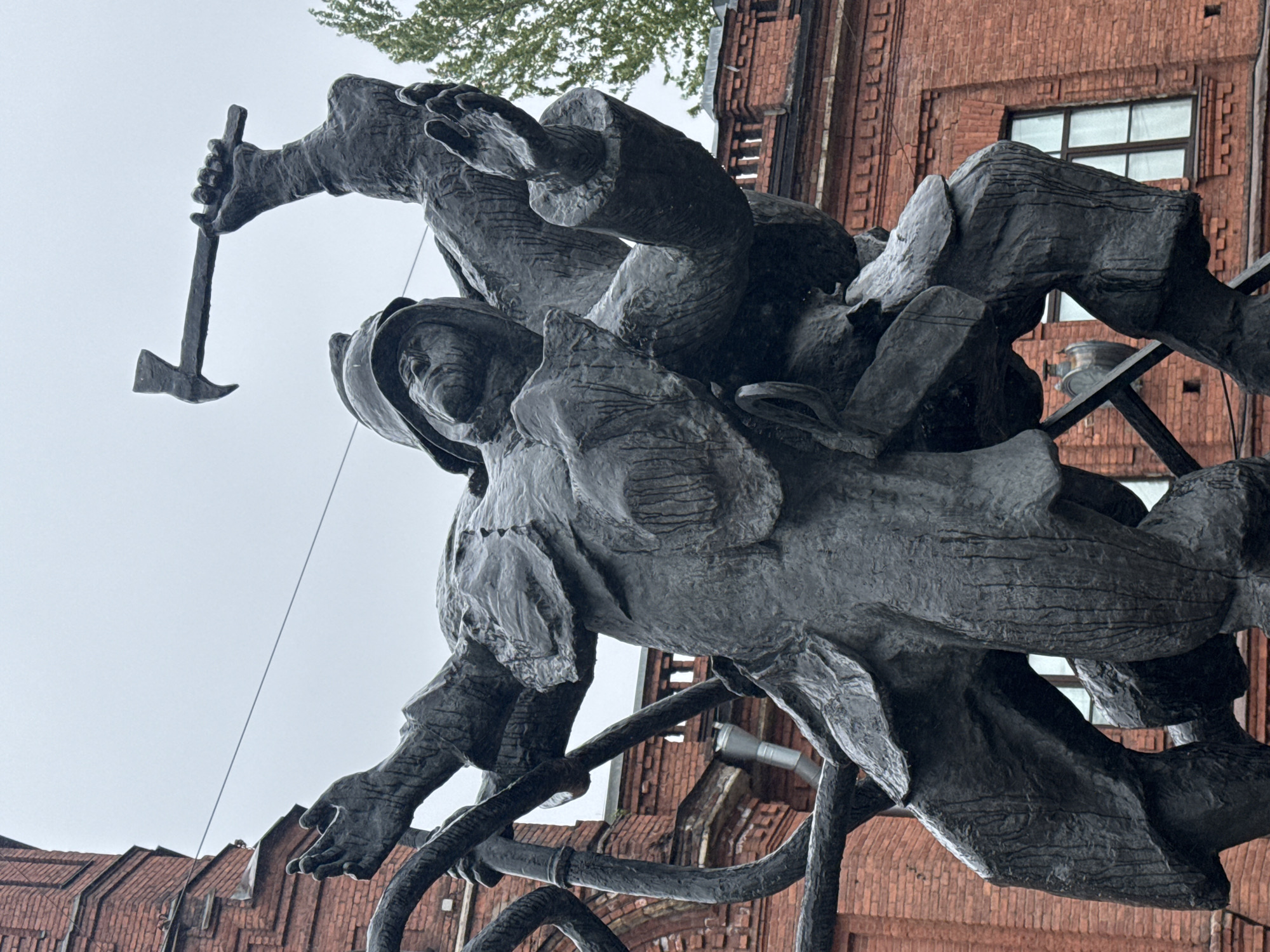 A low-angle photograph captures a dynamic, sculptural group against a partly cloudy sky and a brick building. The sculpture depicts two figures appearing to be in action, possibly firefighters or laborers. One figure, in the upper left, holds a pickaxe. The sculpture itself is dark in tone, and the figures are dressed in work clothes. The brick building behind the sculpture is red and has a window with white frames. The location is Saint Petersburg, Russia.