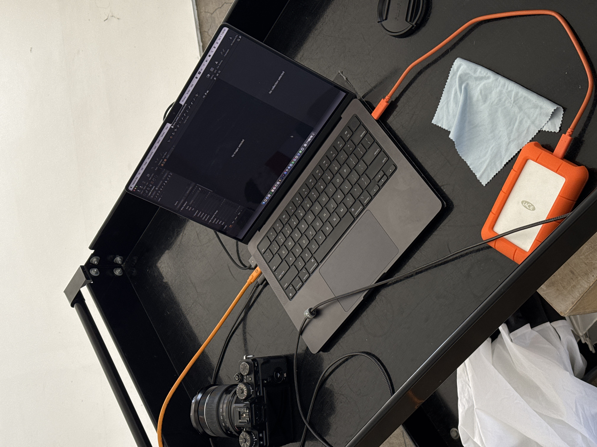 The image shows a workspace on a black table, likely indoors and lit by ambient light. The setup includes a gray laptop with an open screen displaying an image editing interface, a black camera with a lens attached, and an orange external hard drive. An orange cable connects both the hard drive and the camera to the laptop. A light blue cleaning cloth and a black lens cap are also on the table.