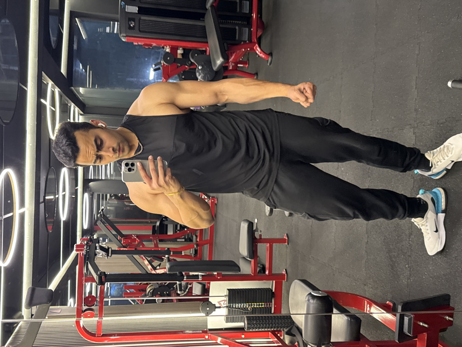 The scene depicts a male individual inside a modern gymnasium located in Kolkata, India. The subject, a man of South Asian appearance with a muscular physique, is centered in the frame, capturing a mirror selfie with a silver smartphone held in his left hand. He is dressed in a black sleeveless top, black track pants, and white athletic shoes with light blue accents. White wireless earbuds are inserted into his ears, and he wears a thin gold-colored chain around his neck and a matching bracelet on his right wrist. His right hand is clenched into a fist. The gym environment features a dark color scheme with black rubber floor tiles, dark walls, and a black ceiling adorned with multiple illuminated circular LED light fixtures. Various pieces of red-framed gym equipment with black upholstery and weight stacks are visible in the foreground, background, and reflected surfaces.