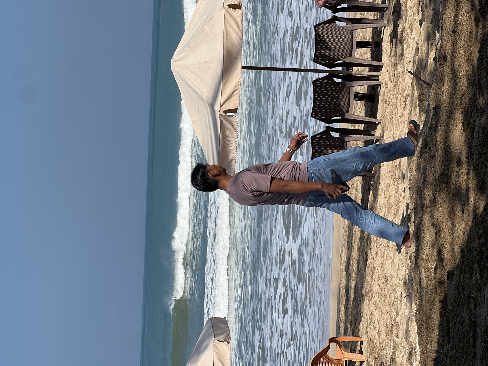 A medium shot captures a man walking on a sandy beach in Kuta, Indonesia, with the ocean and waves in the background. The man, of Asian descent, is wearing a maroon t-shirt and blue jeans, and he is walking from left to right, facing away from the camera and slightly to the left. He is holding a black mobile phone in his right hand. In the foreground, the sand is textured with shadows cast by an unseen object, likely a palm tree. To the left of the man, an orange plastic chair is partially visible. Behind the man, two beach umbrellas are erected, one of which is larger and tan-colored. Several dark brown plastic chairs are arranged in front of the umbrellas. The ocean is calm with gentle waves rolling towards the shore, and the sky is clear and blue. The overall scene suggests a leisurely day at the beach.