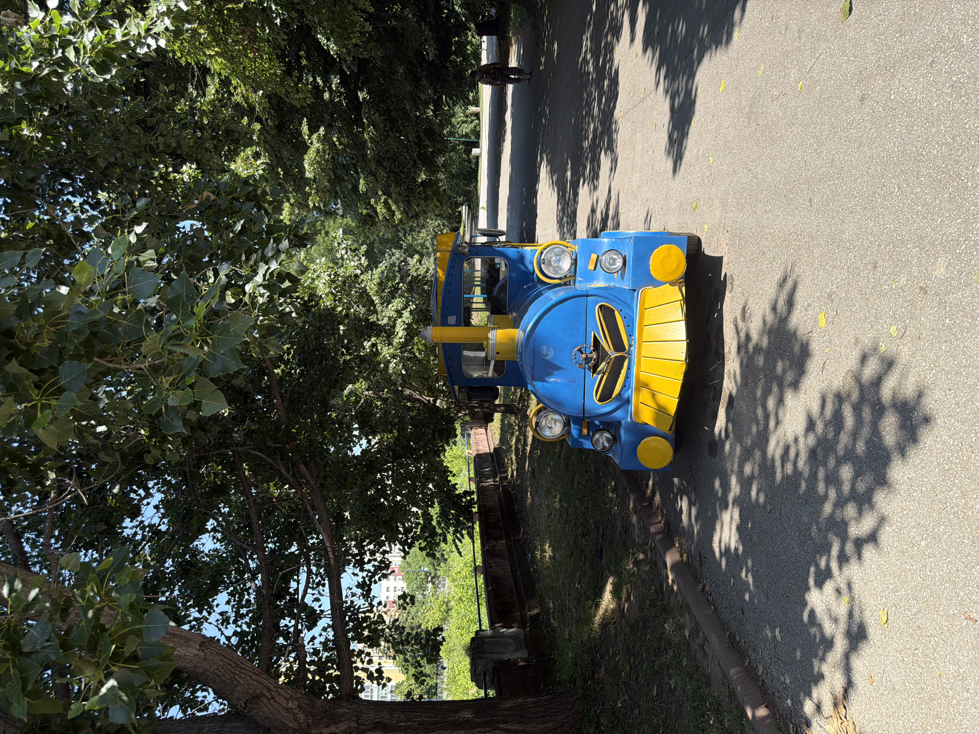 A blue and yellow toy train engine is parked on an asphalt path in a park. The train has a frontal grille and two round headlights. Large trees with green leaves cast shadows on the path. In the background, a person walks away from the camera on the path. Beyond the path, there is a wall and green vegetation. In the far distance, there are apartment buildings. The image is taken from a low angle, looking directly at the front of the train. The location is the City Garden in Pazardzhik, Bulgaria.