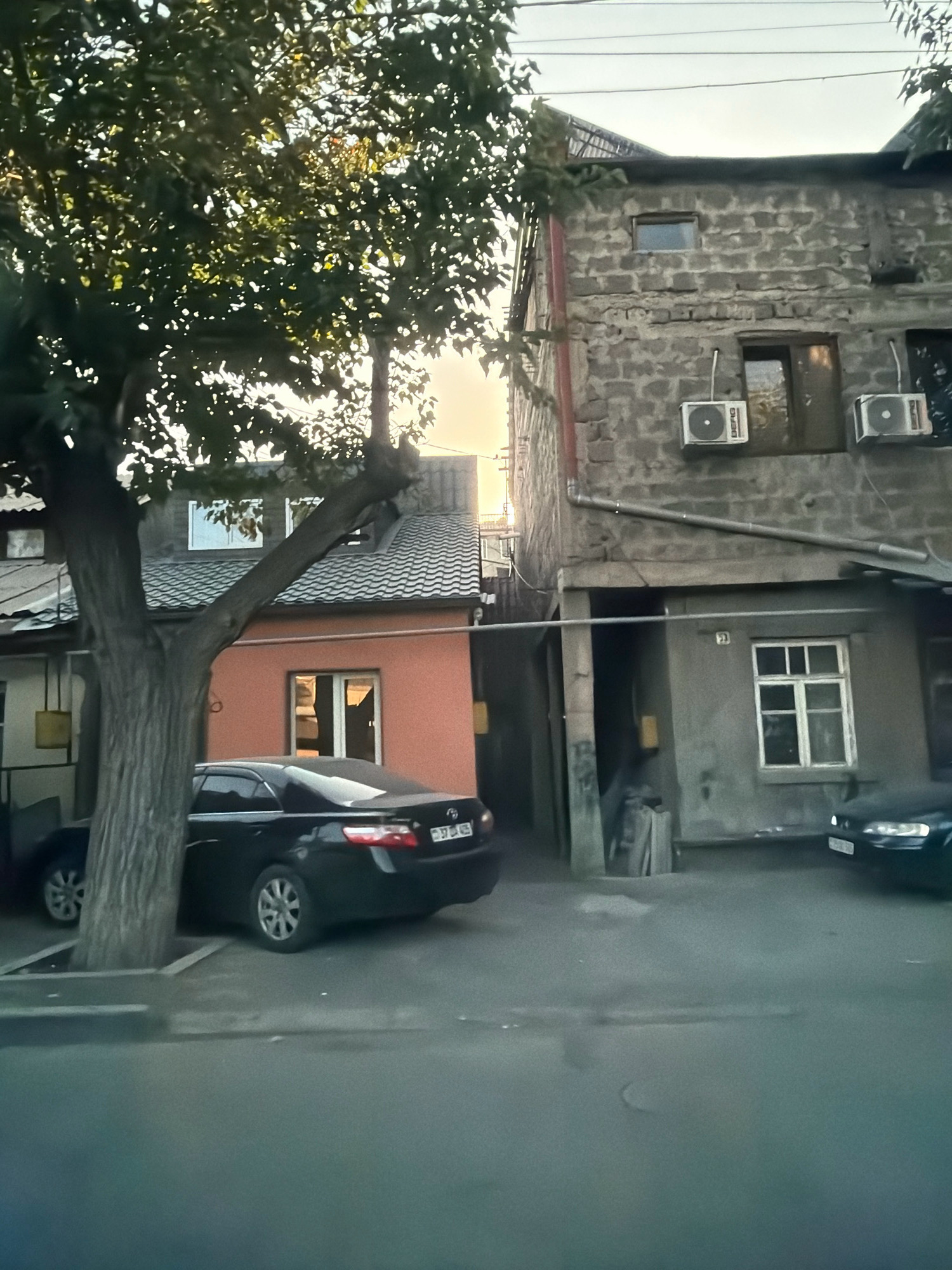 A car is parked in front of a building in Yerevan, Armenia. The building appears to be a multi-story structure with a mix of brick and stucco finishes. There are several windows visible, some with air conditioning units mounted below them. A tree with dense foliage is in the foreground, partially obscuring the view of the building and a black sedan. The sky is overcast with sunlight filtering through the trees.