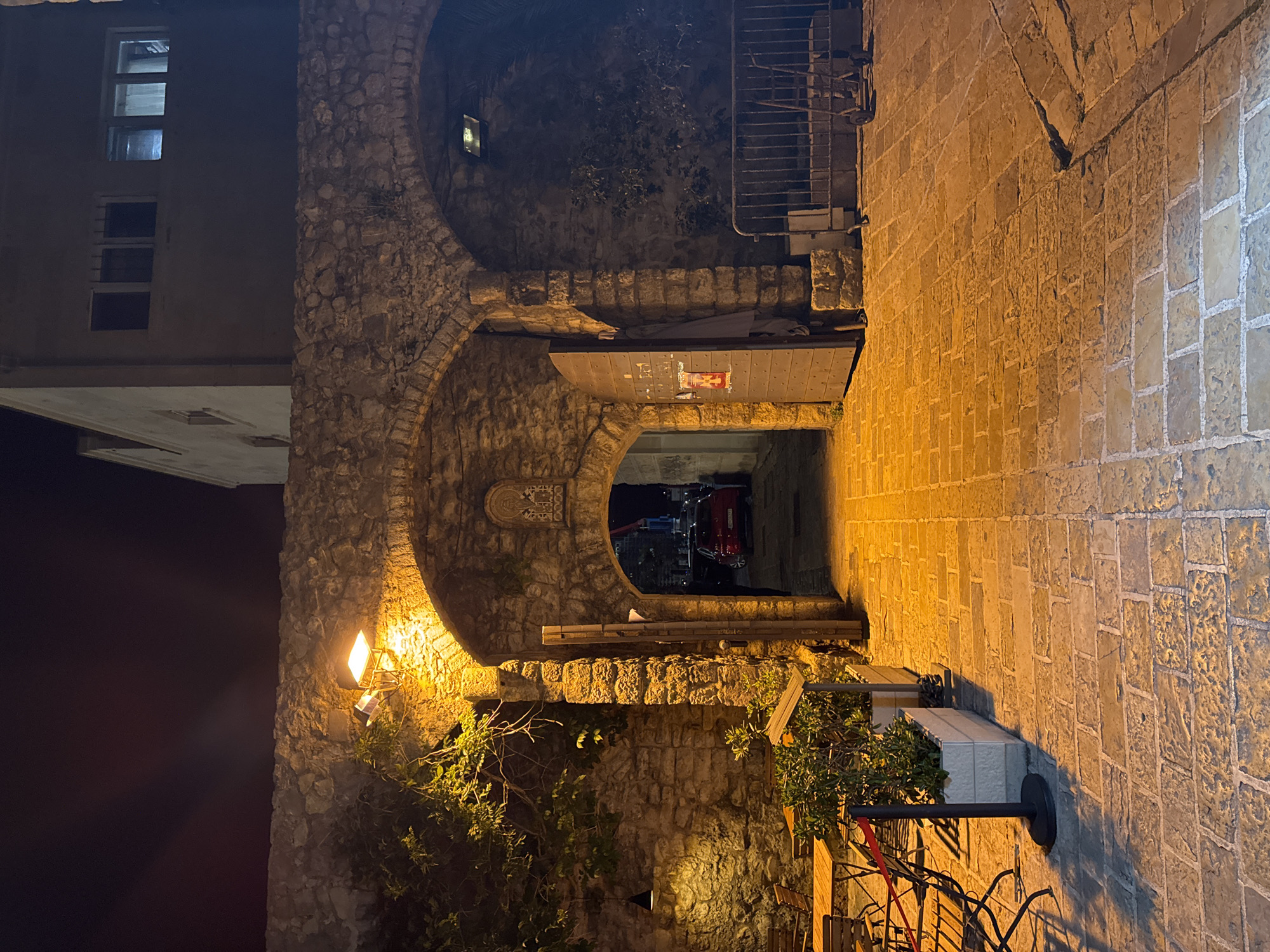 This image captures a narrow, stone-paved street in the Old Town of Budva, Montenegro, at night. The street is lined with stone buildings, including a multi-story building on the left with lit windows. A stone archway forms a passageway, partly covered by a wooden door overhead. Inside the passageway, a red car is visible in the darkness, with streetlights reflecting on the car's surface. A street lamp is affixed to the stone wall near some greenery. An outdoor seating area with metal chairs and potted plants is seen. The light is predominantly warm from the streetlights, creating a contrast with the darkness. The scene is still. No people are present.