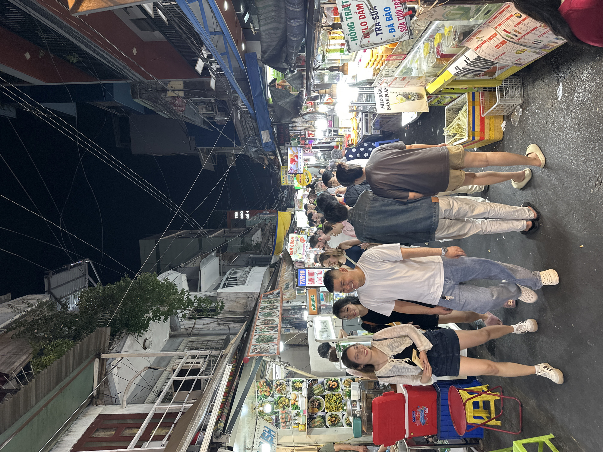 This media file depicts a vibrant night market scene, likely in Thủ Đức, Vietnam, based on the provided context. The image is taken at night, illuminated by numerous artificial lights from stalls and street lamps, creating a bustling atmosphere.
The narrow street is crowded with many people, casually dressed, seen walking, standing, and browsing the various vendors. Some individuals in the foreground are smiling, suggesting a convivial and relaxed mood.
The street is lined with numerous food and drink stalls, displaying an array of items for sale. Visible signs, many in Vietnamese, advertise a variety of offerings including "HỒNG TRÀ T" (Red Tea), "MILO DẦM" (Milo shaved ice/dessert), "TRA SỮA" (Milk Tea), "ĐÁ BÀO" (Shaved Ice), "SỮA TƯƠI TRÊ" (Fresh Milk), and "BÁNH PLAN" (Flan Cake). Other specialized food items like "KEM CUON Thai Lan" (Thai Rolled Ice Cream) and "BÁNH KHỌT VŨNG TÀU" (a type of mini pancake from Vũng Tàu) are also promoted. A red cooler branded "MATSU" is visible on the sidewalk.
A dense network of overhead electrical wires is prominent against the dark night sky, a characteristic feature of many urban landscapes in the region. The buildings bordering the street are multi-story, contributing to the enclosed, lively market environment. The scene captures the essence of a popular local food destination after dark.