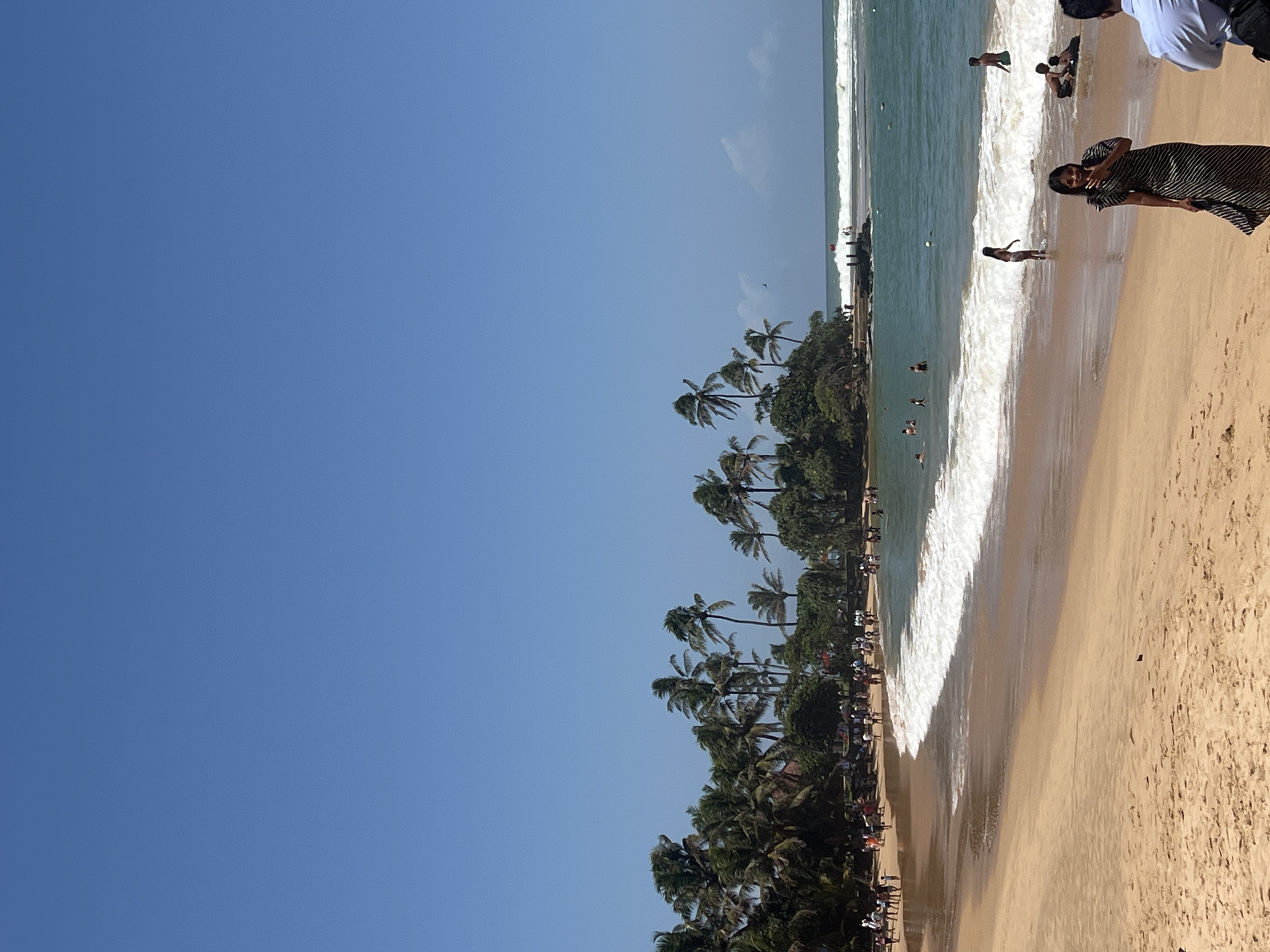 The image depicts a busy beach scene in Hikkaduwa, Sri Lanka, under a clear blue sky. The foreground is dominated by a wide expanse of tan sand, with footprints indicating recent activity. In the mid-ground, a gentle wave is breaking on the shore, with several people wading and swimming in the turquoise ocean. Palm trees and lush greenery line the background, with a rocky outcropping visible in the distance, topped with a small structure. A few people are visible on the sand in the mid-ground and foreground, including a woman in a striped dress looking towards the camera and a man in a light blue shirt in the far right. The overall impression is of a lively tropical beach with a clear, sunny day.