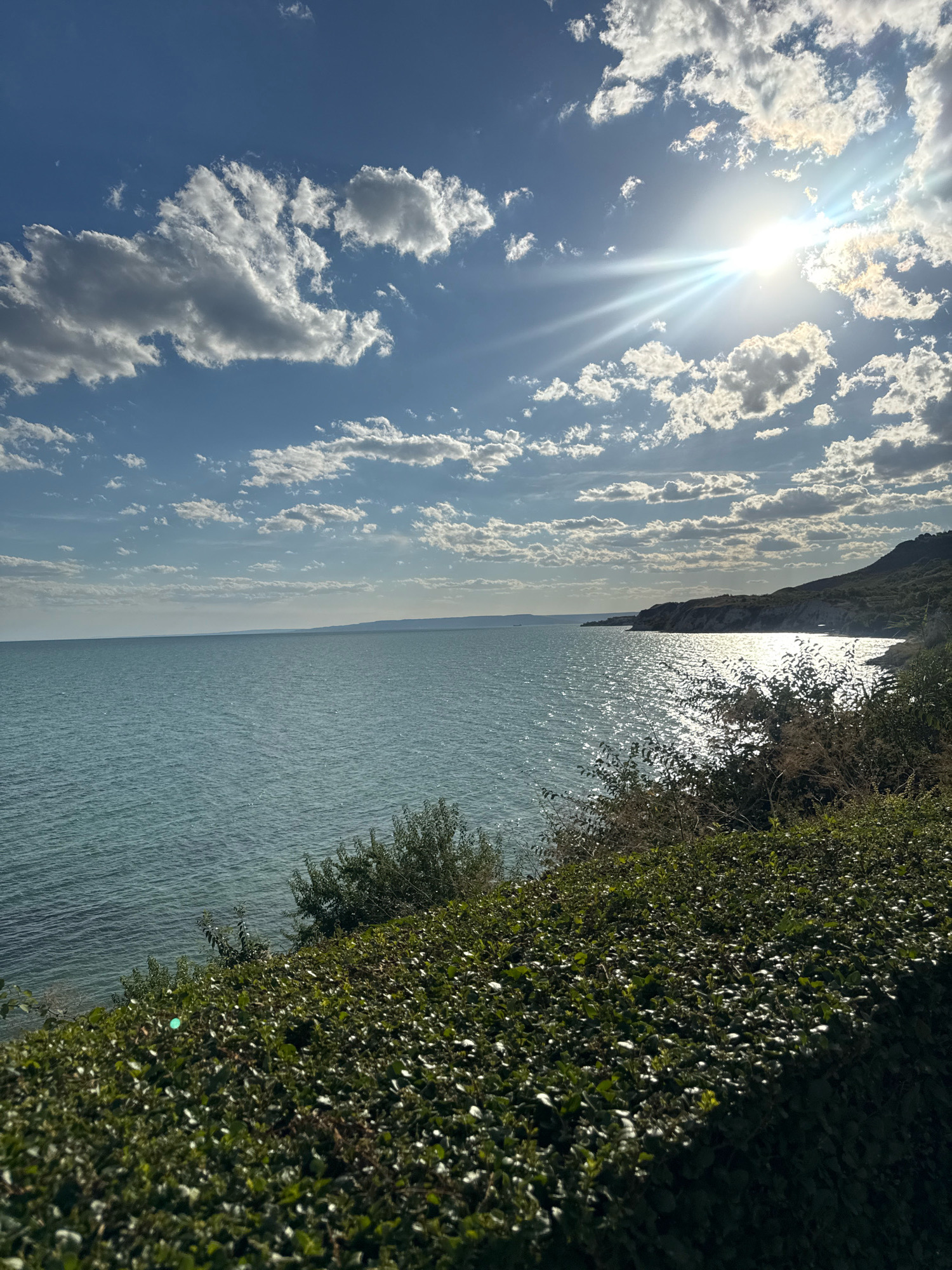 The image shows a coastal view with the sun shining brightly in the sky. The sea is a calm blue-green, with ripples reflecting the sunlight. In the foreground, there is a dense, green hedge. In the middle ground, there are some scattered bushes. The background features a distant coastline with hills and cliffs. The sky is a vibrant blue with scattered white clouds, some of which are illuminated by the sun. The sun itself is visible as a bright starburst with prominent rays. The scene appears to be a scenic overlook of a coastline on a sunny day.
