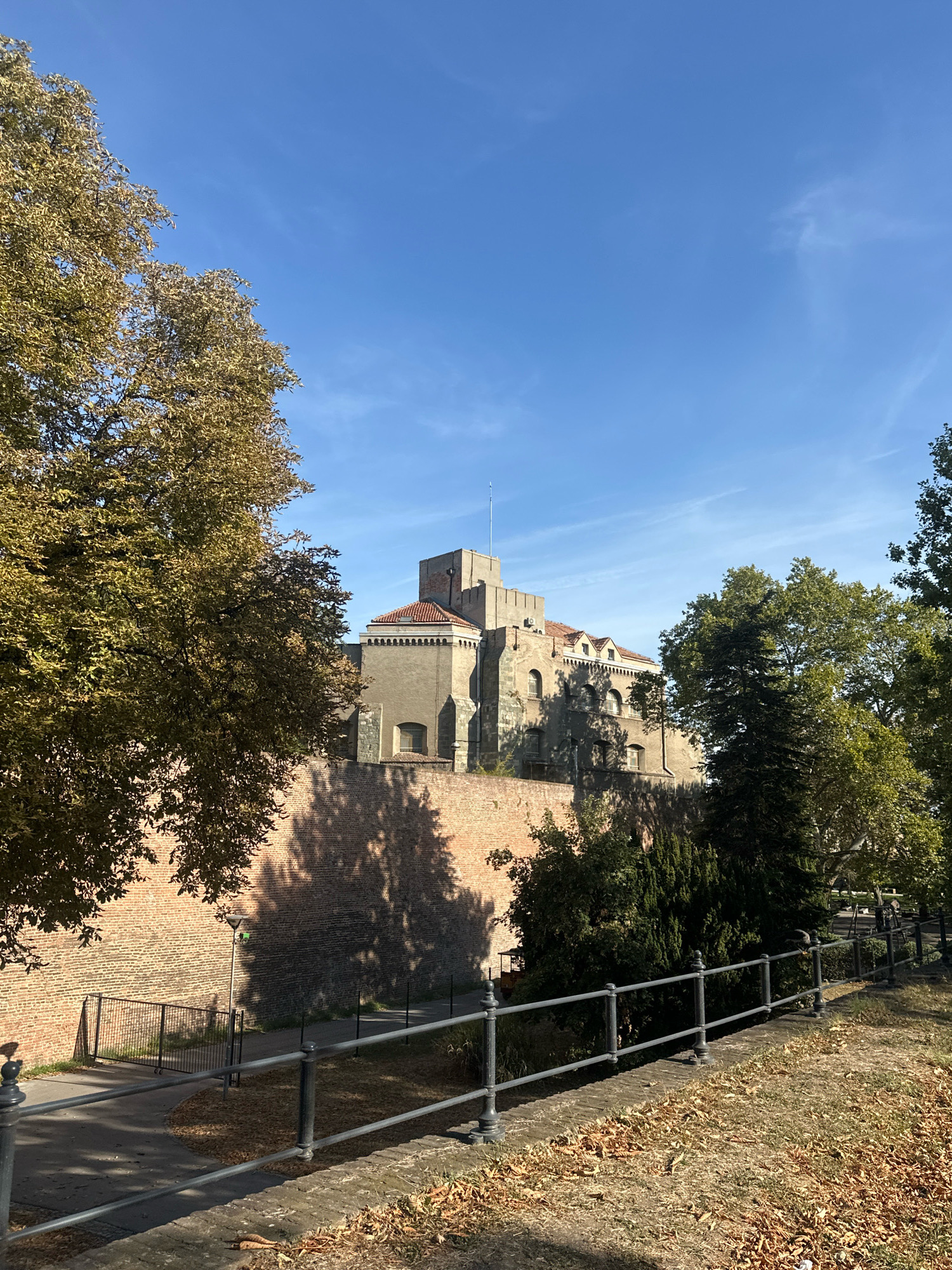 The image shows a view of the Belgrade Fortress, also known as Kalemegdan, in Belgrade, Serbia. The fortress, constructed from brick and stone, is situated on a hill overlooking the confluence of the Sava and Danube rivers. A section of the fortress wall and a building with red-tiled roofs and arched windows are visible. Trees with autumn foliage are in the foreground and to the right, casting shadows on the wall. A paved pathway and a metal railing run along the edge of the hill. The sky is clear blue with a few wispy clouds. The scene appears to be a park or a viewpoint area associated with the fortress. There are no people visible in the image.