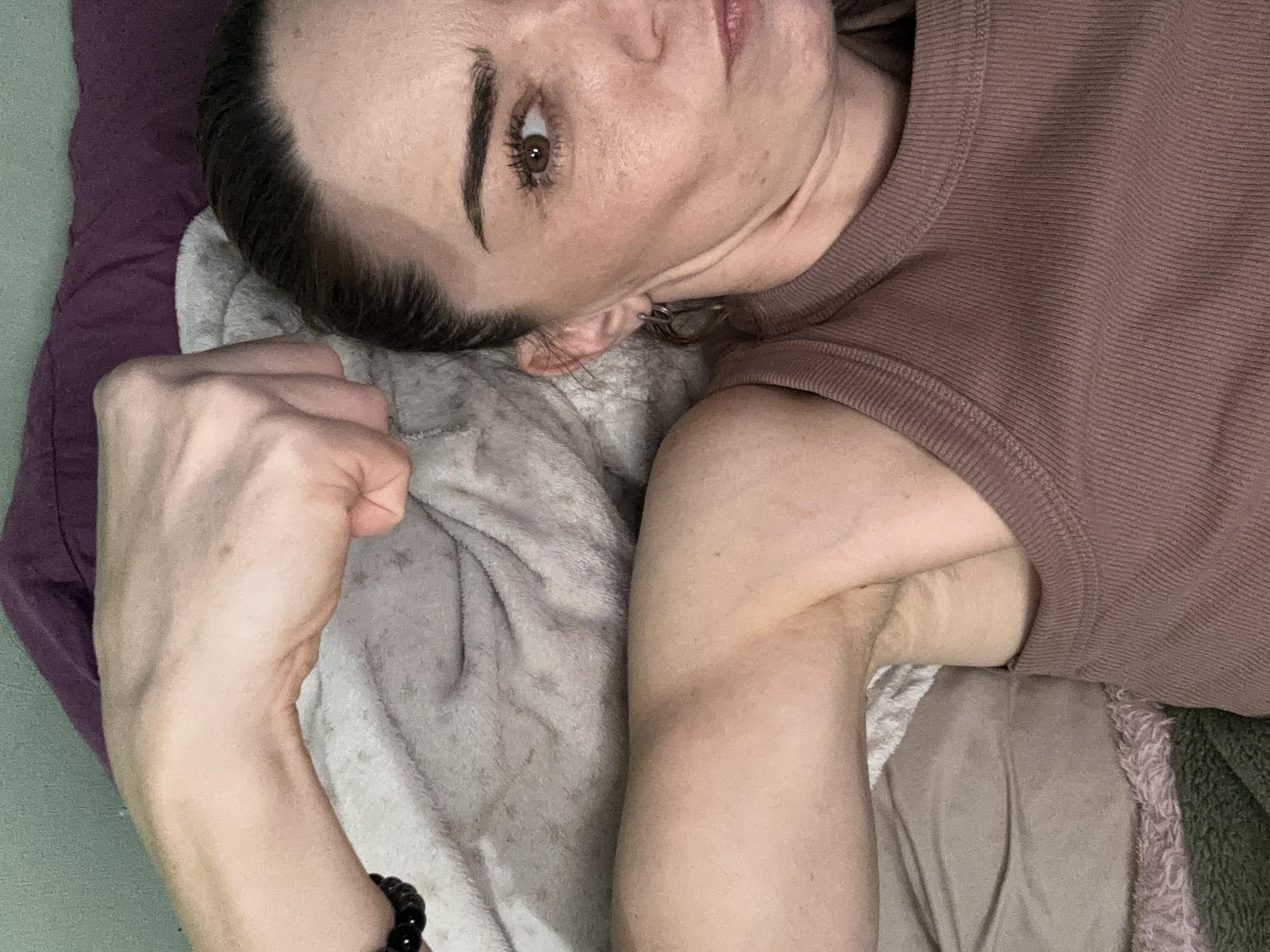 A woman is lying down on a bed, her arm is bent at the elbow and her fist is clenched. She is looking towards the camera with a serious expression. The room is dimly lit, and the bed is covered with blankets and pillows. The woman is wearing a brown tank top. There is a bracelet on her wrist.