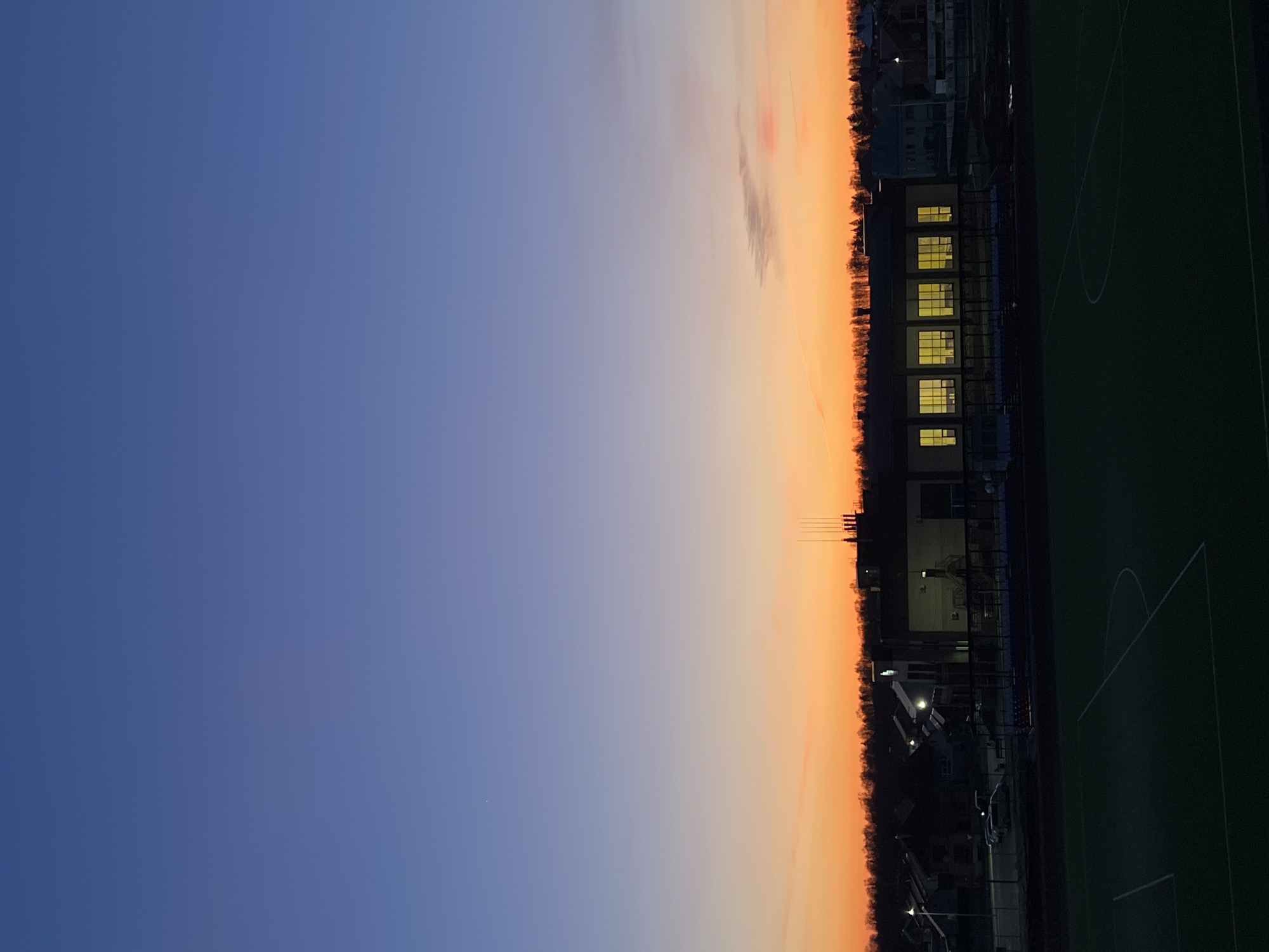 This image captures a twilight scene over a sports complex or school grounds, likely during sunset in Noviy, Russia. The sky dominates the upper portion of the frame, displaying a striking gradient from a deep indigo blue at the top to a vibrant orange and yellow along the horizon. A few faint, wispy clouds are visible in the warmer parts of the sky. In the far upper left, a tiny bright speck, possibly a star or planet, is discernible.
In the foreground to the right, a dark green artificial turf sports field is visible, marked with distinct white lines for a goal area and a partial center circle. No individuals are present on the field. To the left of the field stands a low-rise building with several rectangular windows that are brightly illuminated from within, casting a warm yellow glow. Beyond the building and stretching across the horizon, a dark silhouette of dense trees and distant structures creates a stark contrast against the colorful sky. The artificial lighting from the building and the empty field suggest that the location is ready for evening activities or has just concluded them. No visible text is present in the image.