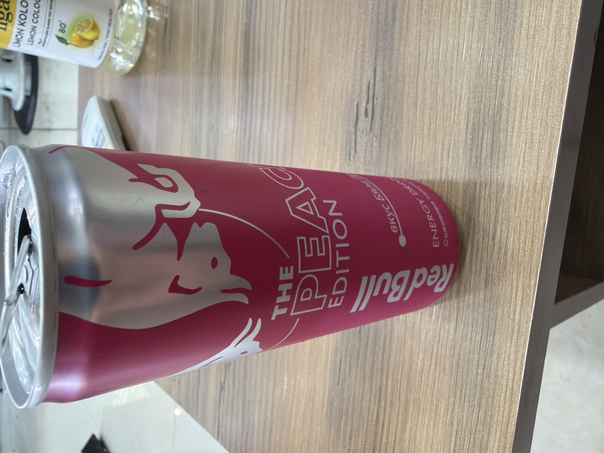 The image shows an open can of Red Bull The Peach Edition on a wooden surface. The can is silver and pink with the brand name and edition printed on the side. To the left of the can, partially visible, is a bottle of "LIMON KOLO/LEMON COLOGNE". The setting appears to be indoors, likely a table surface. The location, Dalgopol, Bulgaria, is inferred as the writing on the Red Bull can includes Bulgarian Cyrillic script.