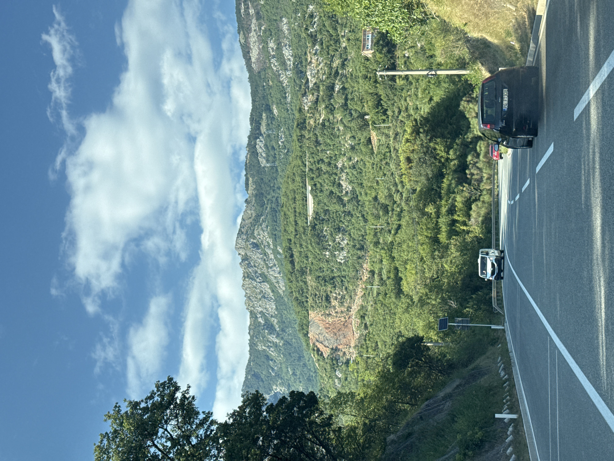The image captures a view from a car driving on a paved road through a mountainous, forested area, possibly near Markovići, Montenegro. The road has clear white lane markings. On the right side of the road, a black Ford Focus with license plate "PG FT938" is in the foreground. Further ahead on the left, a white van is visible. In the background, a steep, tree-covered mountainside rises, with a section of exposed reddish-brown earth near the top. The sky is blue with scattered white clouds. A few utility poles are present along the roadside. The overall scene suggests a scenic drive through a rural, hilly region.