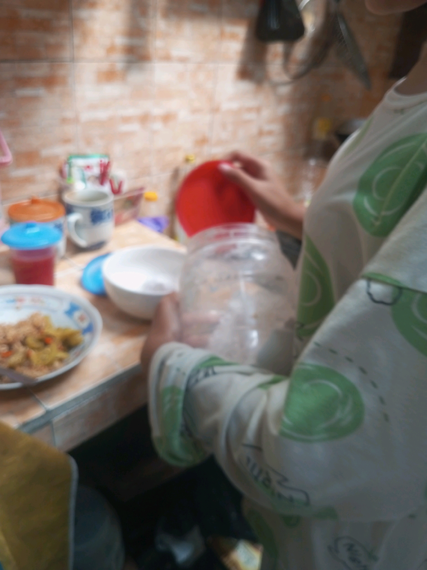 The image depicts a close-up, out-of-focus view of a kitchen scene, likely in Andongsili, Indonesia, given the provided context. The primary subject is a person wearing a white shirt with green circular patterns, holding a clear plastic jar with a white substance inside. Their hands are visible, and they appear to be in the process of adding the substance to a white bowl.
On a tiled countertop, several objects are visible. To the left, a white plate contains a portion of rice with mixed vegetables. Next to it, a blue cup with a blue lid holds a red liquid. Behind the blue cup, an orange-lidded container is partially visible. A white mug with blue intricate patterns sits behind these items, and to its right, a large red bowl is present. In the background, a tiled wall and some kitchen utensils are blurred and indistinct. The overall impression is a domestic kitchen setting during food preparation.