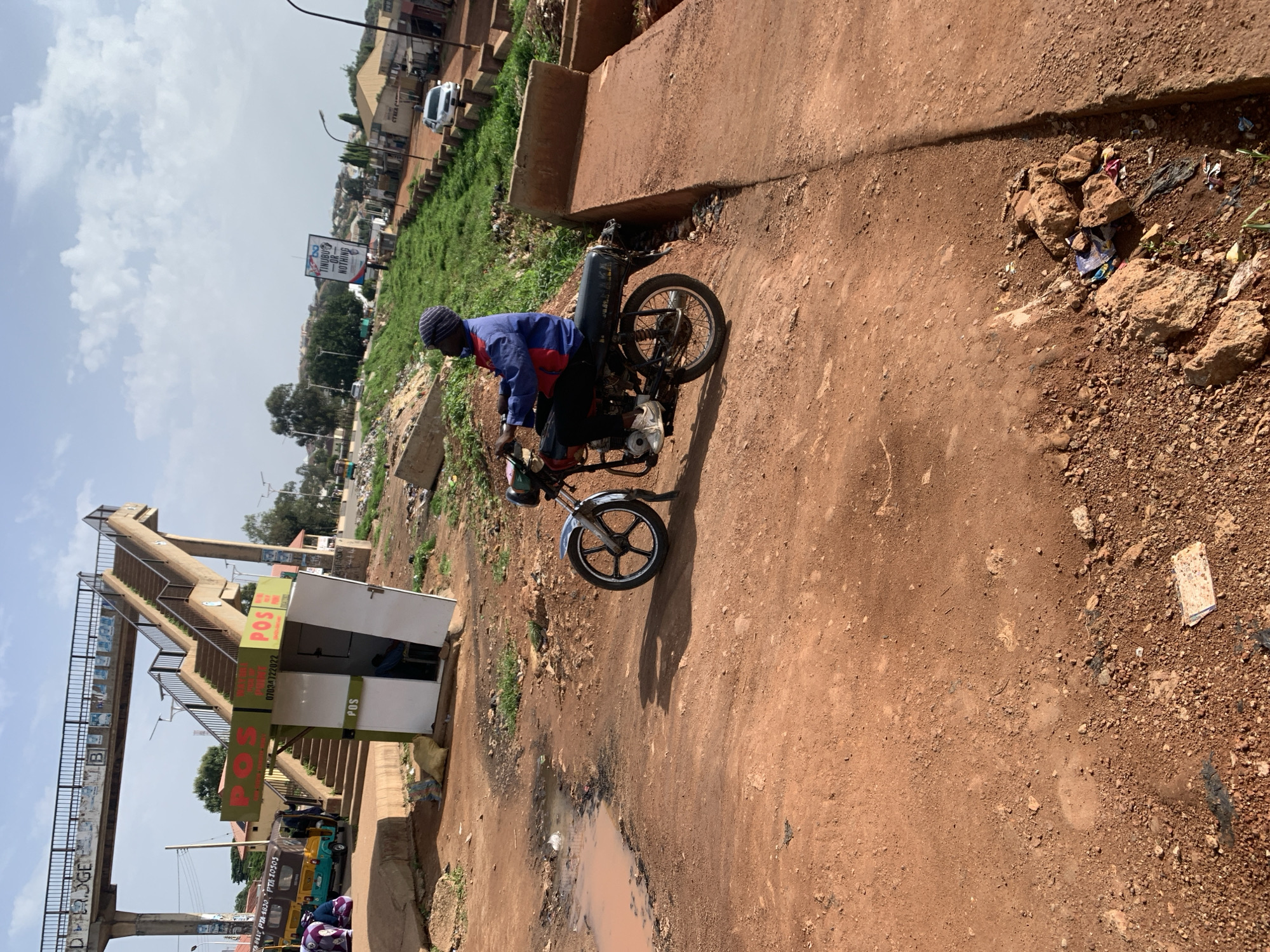 The image captures an outdoor scene in Gura Top, Nigeria, likely a public area with a bridge overhead and a small structure labeled "POS". A person is seated on a motorcycle, facing away from the camera and towards the right side of the frame. The motorcycle is parked on a dirt path, with reddish-brown soil dominating the foreground. Puddles of water are visible on the left, suggesting recent rain or poor drainage. In the background, a pedestrian bridge with stairs is prominent, and a small kiosk with "POS" signage is positioned near its base. A few other vehicles, including a three-wheeled auto-rickshaw, and some people can be seen further in the background near the bridge. The overall environment appears somewhat unkempt, with scattered debris and vegetation along the sides of the path. The sky is partly cloudy.