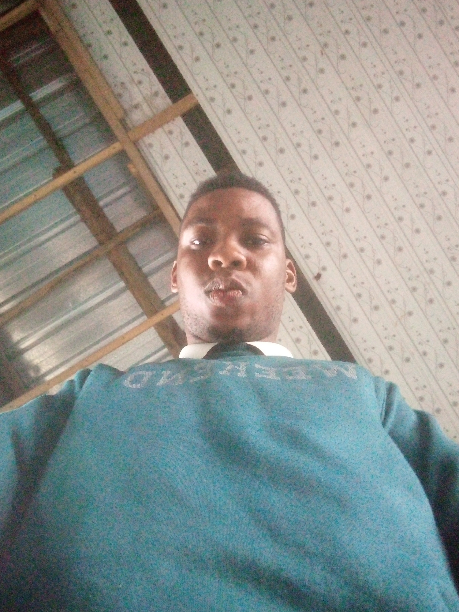 A low-angle, indoor self-portrait shows a dark-skinned man wearing a teal sweater with a white collar shirt underneath. The upper part of his body is centered in the frame. The background includes a ceiling constructed of multiple materials: metal sheeting with visible frame, and white patterned tiles. Based on the ceiling construction, the location is most likely a residential home. There is no other information to confirm the location in the city of Ikorodu, Nigeria.