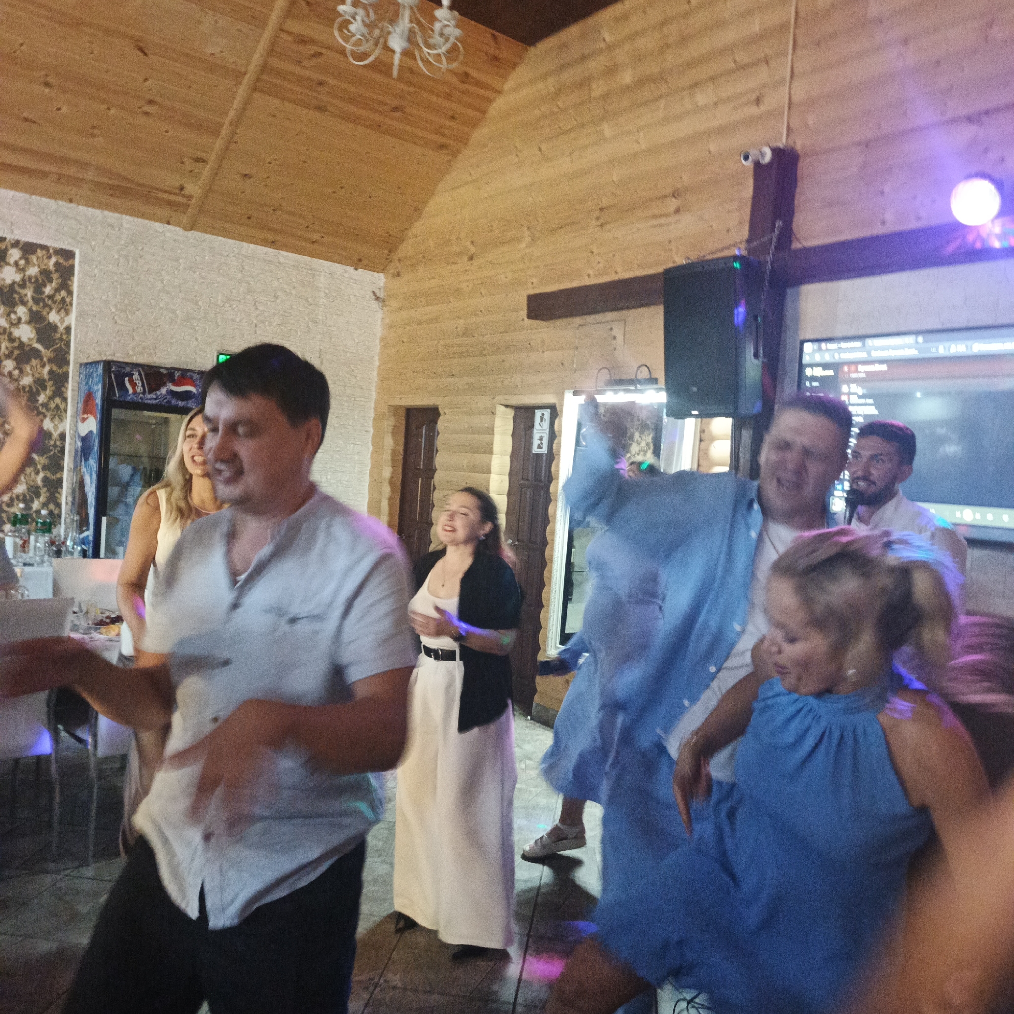 In the interior of a wooden building, a group of people are dancing and enjoying themselves. The scene appears to be a party or celebration. In the foreground, a man in a light-colored shirt and dark pants is actively dancing, his arms in motion. To his left, another person's arm is visible. Behind him, a woman with blonde hair in a white dress is smiling and looking towards the side. A woman in a long white dress and a black jacket is also visible, facing forward with her hands together. Further back, a man in a light shirt is holding a microphone and appears to be singing or speaking. A woman in a blue dress is in the right foreground, also dancing. The environment includes a wooden ceiling and walls, a Pepsi refrigerator on the left, and a large screen displaying content on the right. Colored lights are present, indicating a festive atmosphere. The specific location in the city of Chaikovsky, Russia, is not discernible from the image itself, but the overall scene depicts a social gathering.