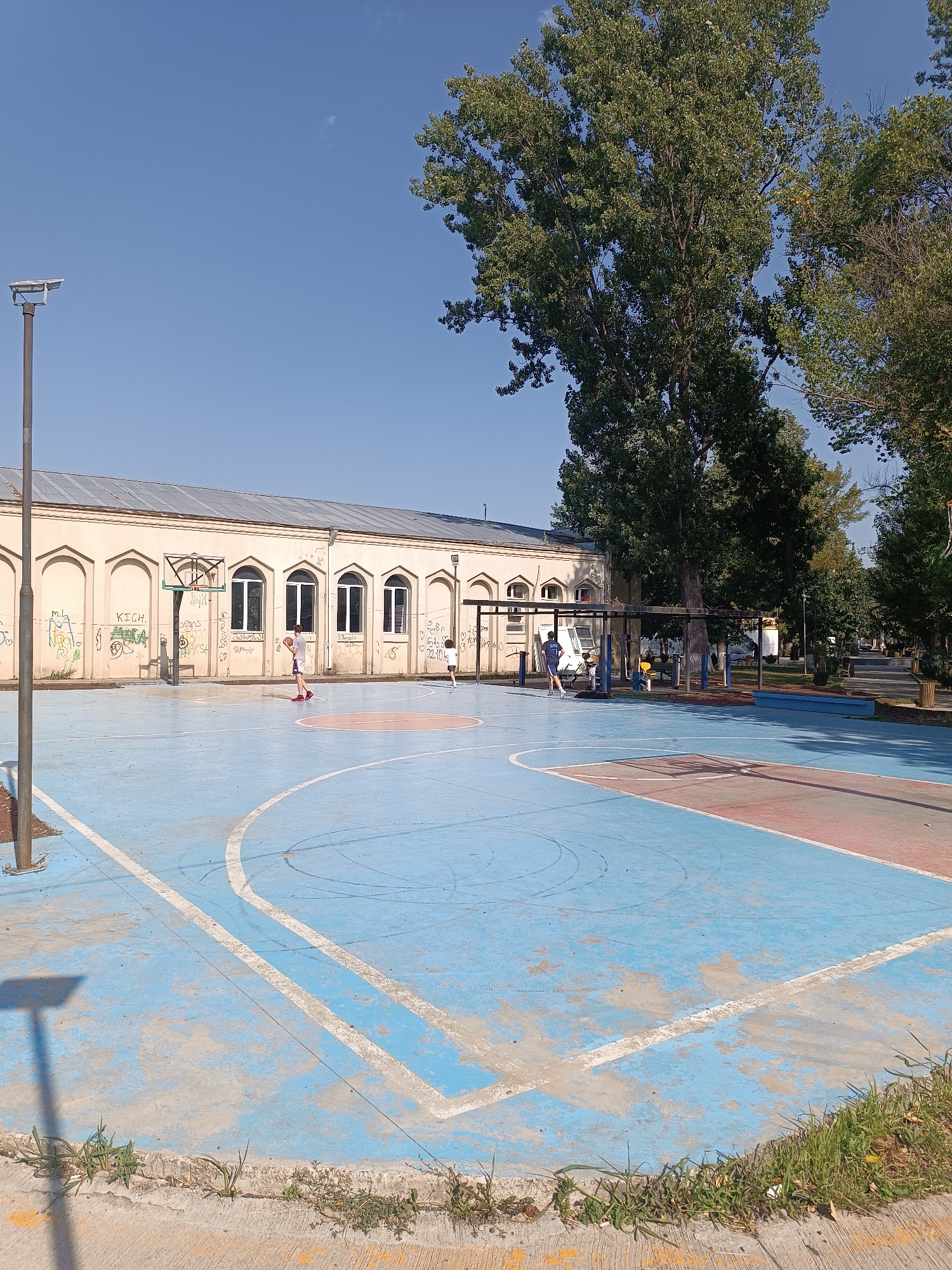 This image depicts an outdoor basketball court in the city of Khashuri, Georgia. The court is painted blue with red areas, marked with white lines. A single basketball hoop is visible on the left side of the court. Two individuals are present on the court: one person is standing near the hoop, appearing to dribble a basketball, while another person is on the right side of the court, walking or running.
In the background, a light-colored building with arched windows and a metal roof is situated. The building is covered in graffiti. Adjacent to the building and behind the court are several large, mature trees with green foliage, suggesting a park-like setting. A street light with a solar panel is positioned on a pole to the left of the court. The sky is clear and blue, indicating fair weather. The overall scene suggests a public recreational area.