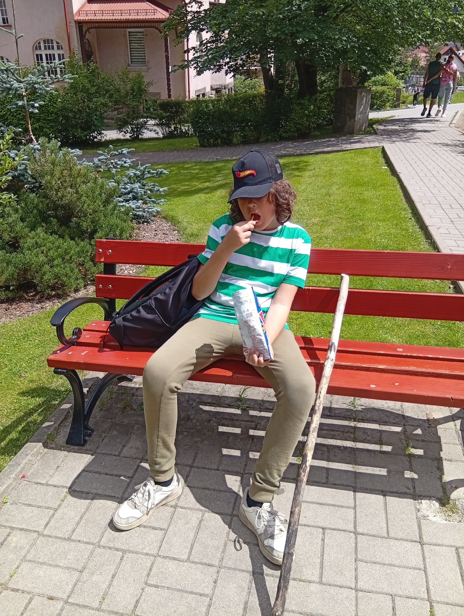 In Karpacz, Poland, a young person wearing a green and white striped t-shirt, khaki pants, a black baseball cap, and white sneakers sits on a red park bench. The person is holding a bag of chips and eating from it, with a stick propped up on the bench next to them. A black backpack is on the bench to their left. The scene is set outdoors with a building in the background, trees, and a grassy area with a paved walkway. Two people are visible walking on the walkway in the background.
