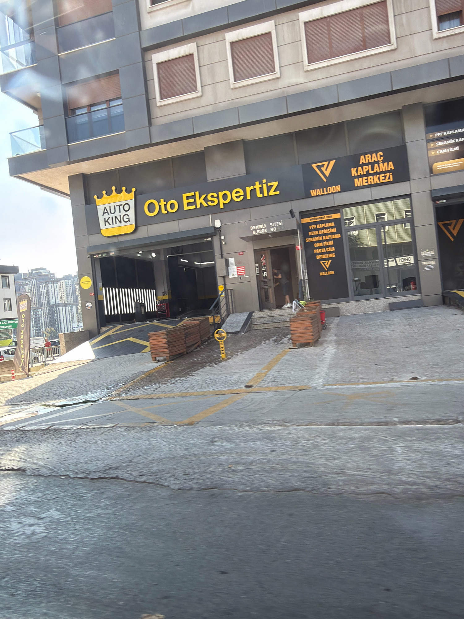 The image shows a storefront for "AUTO KING" and "WALLOON" on a sunny day in Ataşehir, Turkey. The AUTO KING store appears to be a car service or detailing business, with a large sign featuring a crown logo. Next to it, WALLOON is advertised as an "ARAC KAPLAMA MERKEZI," which translates to "Car Coating Center." This section lists services such as PPF kaplama (paint protection film), seramik kaplama (ceramic coating), cam filmi (window film), and pasta cila detailing (polishing and waxing).
In front of the building, there is a ramp leading into the service bays, marked with yellow lines on the asphalt. Several wooden crates are stacked near the entrance. A person is partially visible entering the building. In the background, modern apartment buildings are visible, indicating an urban setting. A banner with Turkish text and a logo is displayed on a pole to the left of the AUTO KING entrance. The ground in the foreground appears wet, possibly from recent rain or cleaning.
