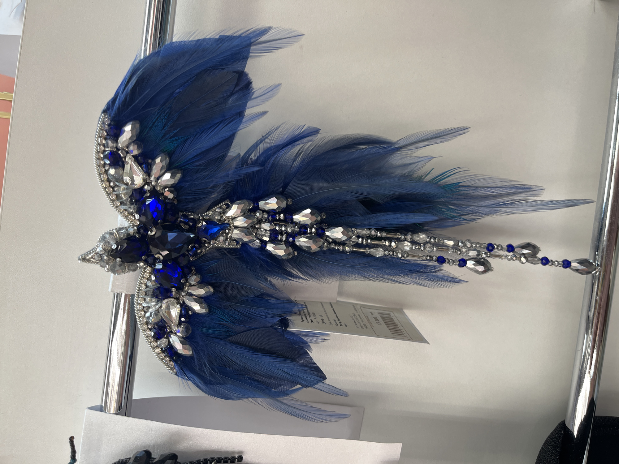 The image shows a close-up of a decorative bird-shaped brooch with blue feathers and sparkling rhinestones and blue gemstones. The brooch is hanging on a metal rod, likely in a retail setting. A tag with text in Russian is attached to the brooch. The background is a plain white wall. The location of this scene is in the city of Svetly, Russia, as indicated by the text on the tag which includes Cyrillic script and a price in Russian currency.