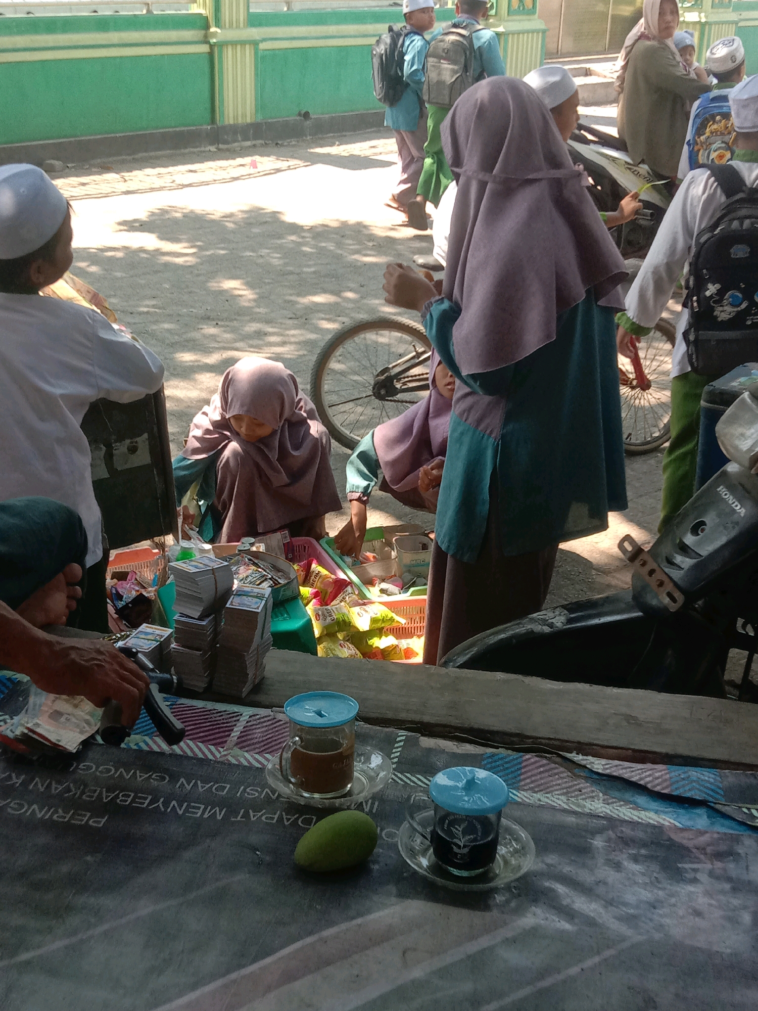 In Ngempit, Indonesia, on a sunny day, a group of people are gathered near a green and yellow striped wall. In the foreground, a person's hand is seen reaching for what appears to be money and small packages. Two glasses of drinks, one with a blue lid, sit on a table with a printed cloth. Next to the drinks, a green mango rests on the table. Behind the table, a woman in a purple hijab crouches, surrounded by various items including stacks of cards and bags of snacks. A motorcycle is partially visible on the right side of the image. In the background, several individuals wearing religious attire and backpacks are walking. The ground is covered in gravel and sand, with shadows cast by the bright sunlight.