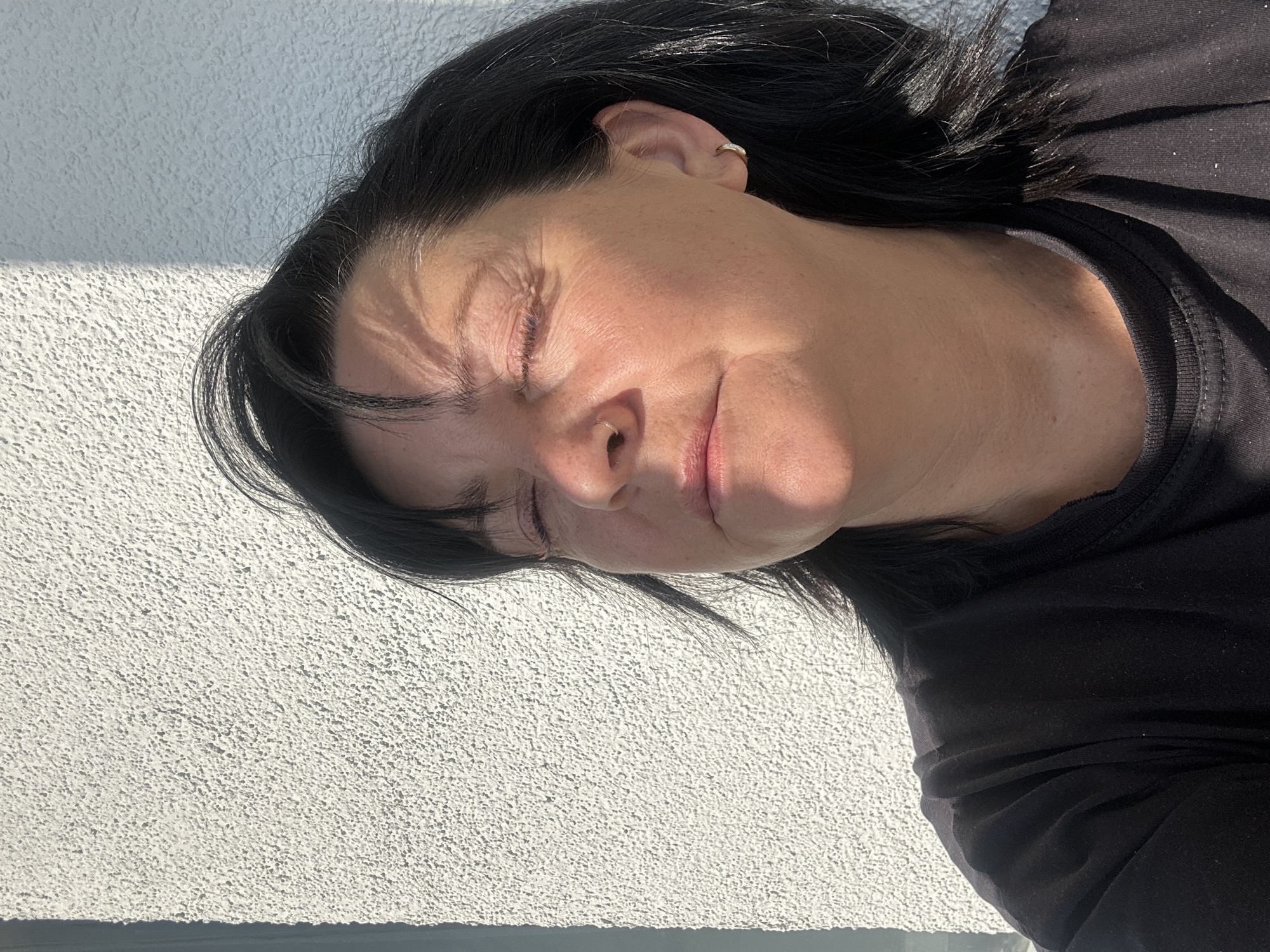 A woman with dark hair lies with her eyes closed in an outdoor setting. Sunlight casts patterns on her face. She wears a black top. The background shows a textured, light-colored wall. There is no indication of a city or Dnp Ryzhiki, Russia.