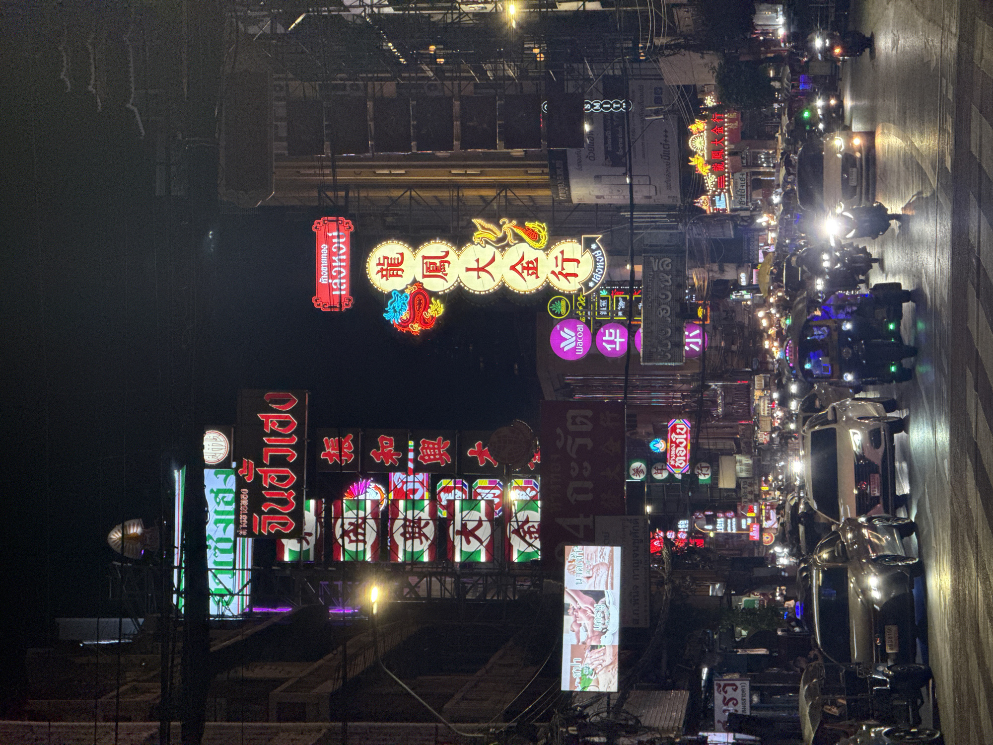 This image depicts a bustling night scene on Yaowarat Road (Chinatown) in Bangkok, Thailand. The street is heavily illuminated by a multitude of neon and LED signs, predominantly featuring text in both Thai and Chinese script.
Prominent signage advertises gold shops, identified by names such as "เล่งหงวน" (Leng Hong) and "จินฮั่วเฮง" (Chin Hua Heng), with one vertical sign for "เล่งหงวน" featuring a stylized dragon and phoenix. Other commercial signs include "สปา นวดแผนไทย" (Spa Thai Massage) and the brand "Wacoal." The architecture consists of multi-story commercial buildings lining the thoroughfare, their facades largely covered by the dense array of brightly colored signs. A faint, traditional Chinese-style tiered pagoda roofline is discernible against the dark sky in the upper right background. Overhead, a complex network of utility cables crisscrosses the street.
The road is occupied by moderate to heavy vehicular traffic. Numerous cars, predominantly sedans and SUVs, travel with their headlights illuminated, reflecting off the damp-looking asphalt. Several motorcycles with riders and passengers are present, weaving through traffic with their headlights and taillights glowing. Multiple three-wheeled tuk-tuks are also visible, particularly in the foreground and along the left side of the road. People, including drivers, riders, and a few pedestrians, contribute to the active urban environment. The scene is characterized by its vibrant artificial light and constant motion.