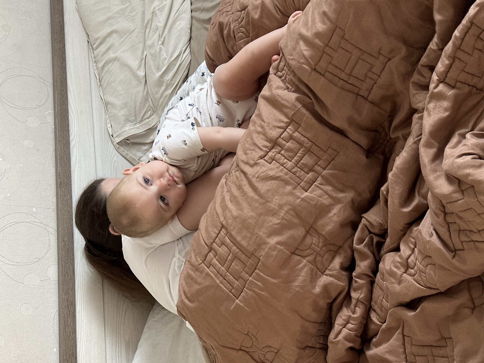 A baby with fair skin and blue eyes is lying on a brown quilted blanket. The baby is wearing a white onesie with a pattern of small cars. The baby is positioned on its side, with its head resting on the arm of an adult who is partially visible behind the baby. The adult has dark hair tied in a ponytail and is wearing a white t-shirt. The background shows a bed with light-colored bedding and a headboard. The overall scene appears to be indoors, likely in a bedroom. The location is Fredenbeck, Germany.