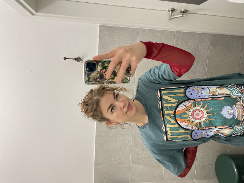 An indoor mirror selfie captures a young adult female from the chest up, positioned slightly left of center. She has light brown, curly hair styled in an updo and light-colored eyes. Her attire includes a teal short-sleeved t-shirt featuring a mirrored "INFINITE" graphic with celestial and feline motifs, layered over a long-sleeved, sheer red mesh top. Her right hand, holding a smartphone with a multi-colored clear case, is extended into the frame to capture the image. She maintains eye contact with the camera, exhibiting a subtle smile. The background consists of a wall divided horizontally, with light grey rectangular tiles on the lower half and a plain white surface on the upper half. A dark, ornate metal wall hook is mounted on the white wall section to the subject's right. A white door with a silver handle is partially visible on the far right. A dark green cylindrical bin is present in the lower-left corner of the frame. The precise location within Budva, Montenegro, is not discernible from the visual evidence, but the environment suggests a functional indoor space, such as a restroom or changing room.