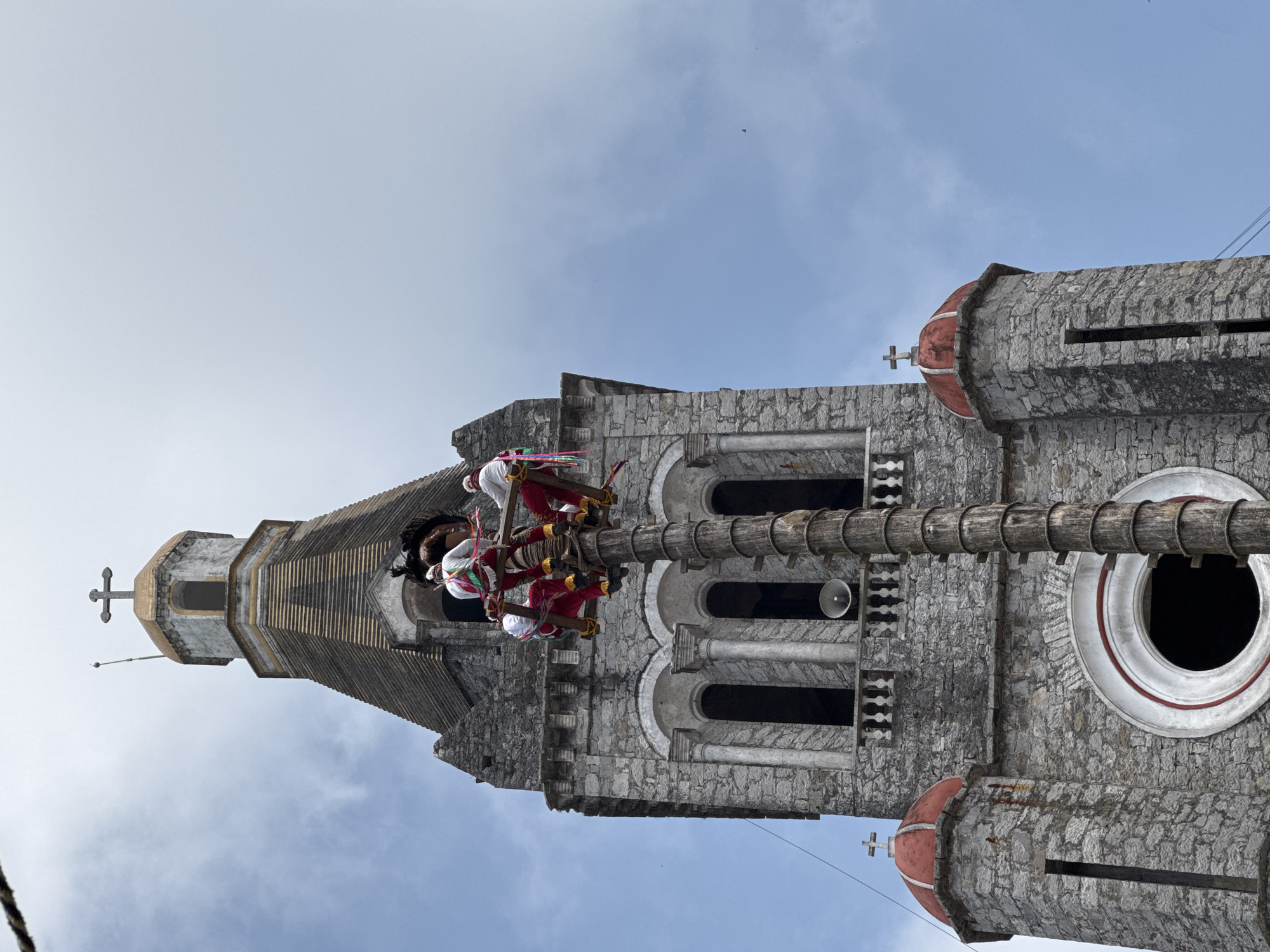 The image shows the Papantla Flyers performing their ritual on a tall pole erected in front of the Iglesia de San Francisco de Asís in Cuetzalan, Puebla, Mexico. Four men in red and white ceremonial outfits are attached to ropes and suspended from the top of the pole. They are in the process of descending in a spiraling motion, symbolizing the descent of birds from the sky. The stone church is visible in the background, with its tower reaching towards the sky. The sky is a light blue, partly cloudy.