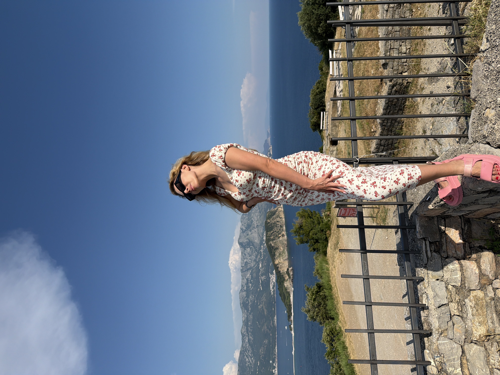 A blonde woman wearing sunglasses, a floral print dress, and pink platform sandals stands on a stone wall overlooking the Adriatic Sea. She is positioned on the right side of the frame, with her left hand on her hip and her right arm resting on her leg. Behind her, the blue sea stretches to the horizon, with mountains in the distance. The scene appears to be taken from the citadel overlooking Budva, Montenegro, with the island of Sveti Stefan visible in the distance. The sky is clear blue with a few wispy clouds. A black metal fence runs along the edge of the wall in the foreground.