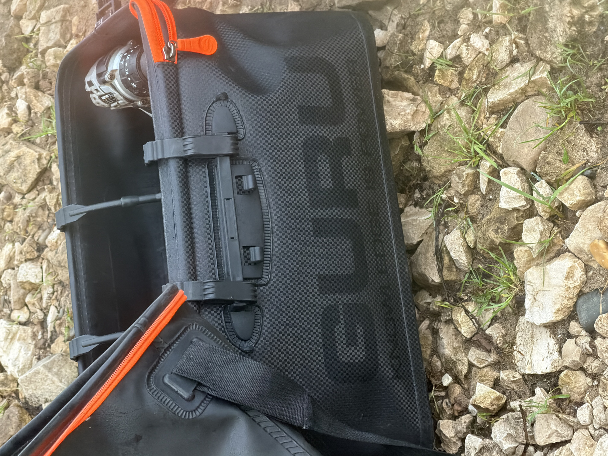 This image presents a black, robust-looking storage case or bag, likely designed for outdoor gear, resting on rocky ground. The case features a prominent carbon fiber-like checkered texture and is highlighted by vibrant orange zippers and matching rubber pull tabs. An orange handle is partially visible at the top edge of the bag. Inside the open case, a silver and black fishing reel is secured to a fishing rod by black plastic clasps and straps. The reel displays numerical markings such as "16," "18," and "20." A subtly embossed brand name, appearing to spell "DAIWA," is visible on the textured surface of the bag. The surrounding ground consists of numerous light-colored, irregularly shaped rocks and pebbles, interspersed with small patches of green grass, indicative of a natural, rugged outdoor setting. The lighting suggests it is daytime, with soft, ambient illumination. The scene depicts fishing equipment either stored or being prepared for use in its protective container. The activity suggests a fishing-related context, set outdoors in Noviy, Russia. No individuals are present, and no active event is depicted beyond the state of the equipment.