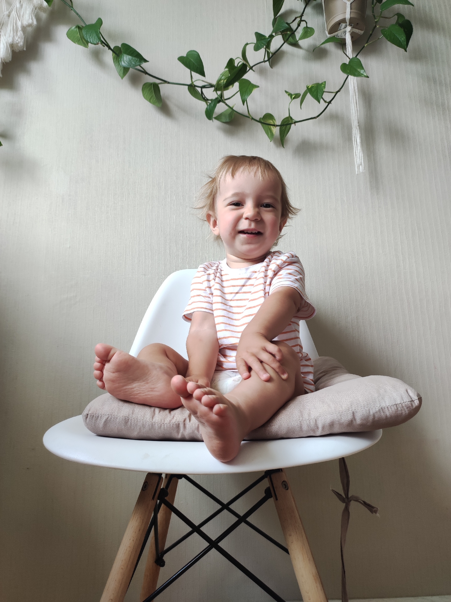 A young child with blonde hair and a striped shirt sits on a white chair with wooden legs, which is placed on a light-colored pillow. The child is smiling at the camera. Green vines with heart-shaped leaves hang from the wall behind the child. The wall has a textured finish. The image appears to be taken indoors.