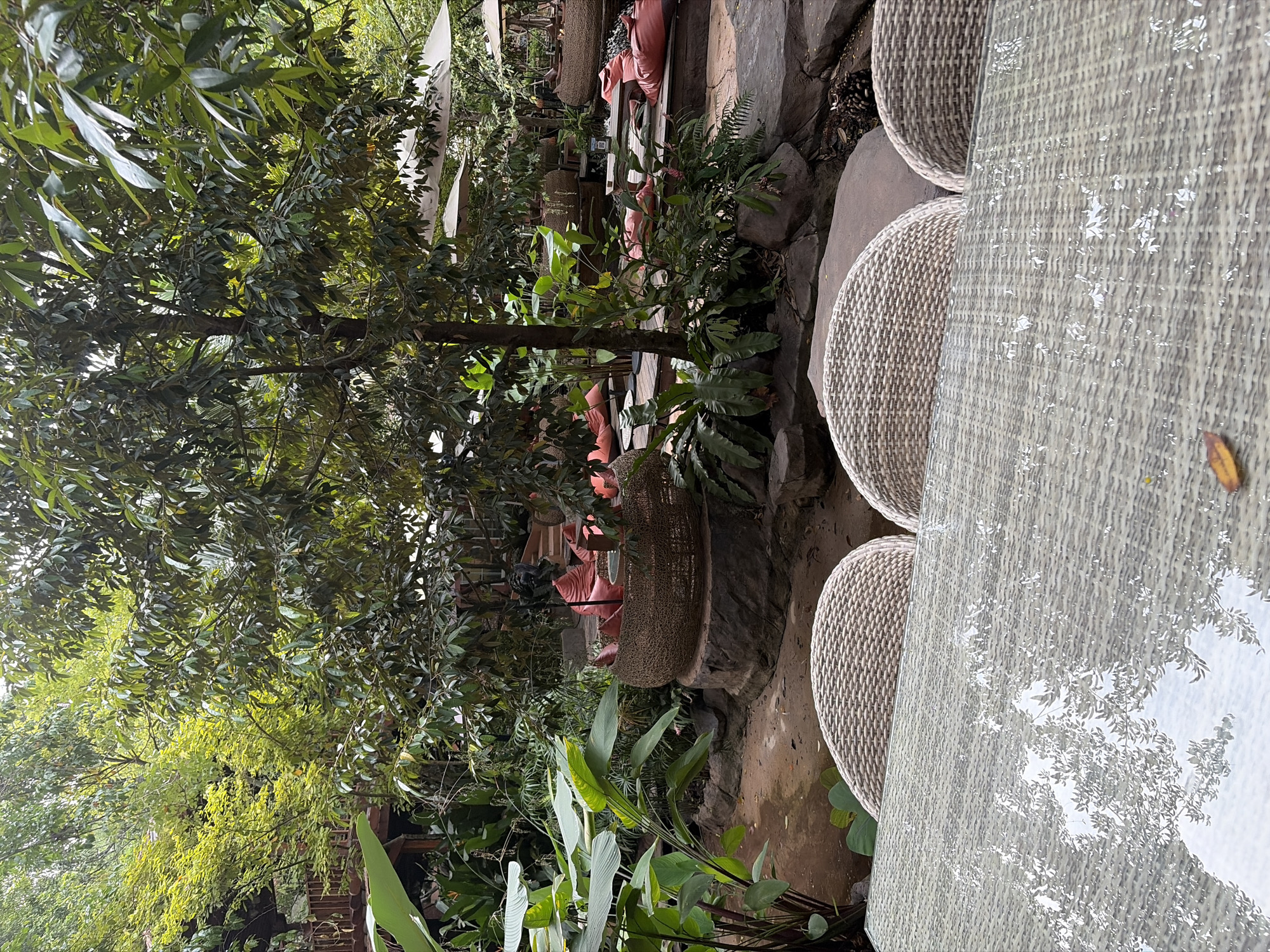 The image shows an outdoor seating area at a cafe or restaurant in Tambon Mae Raem, Thailand. In the foreground, a glass-topped table with a wicker base reflects the surrounding greenery and sky. Three wicker chairs are visible behind the table. The middle ground features a natural landscape with lush vegetation, large rocks, and pathways. Several seating arrangements are scattered throughout the scene, including low wicker tables with large, round cushions in a coral color, and larger, woven basket-like seating structures filled with similar cushions. In the background, trees and foliage create a dense canopy, with glimpses of umbrellas and possibly other seating areas. The overall atmosphere is relaxed and natural, typical of an outdoor dining establishment in a tropical setting.