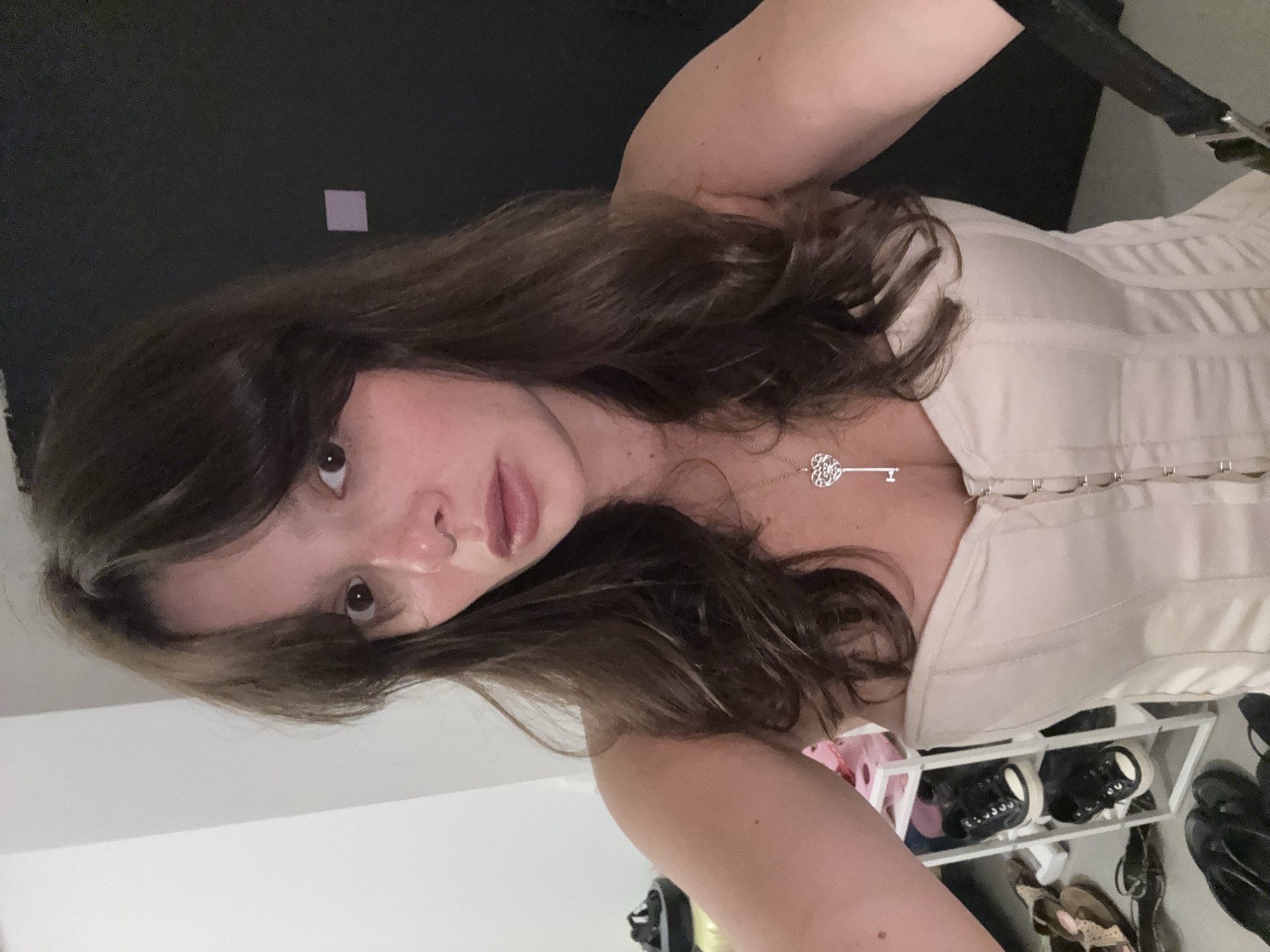 The image shows a young woman from the chest up, posing for a selfie. She has long, dark brown wavy hair and is wearing a light-colored corset with a silver key-shaped pendant. The background is dark and appears to be indoors, possibly a closet or a dressing room, with a shelf of shoes visible to the left. There is no indication of the location being Tel-Aviv, Israel.