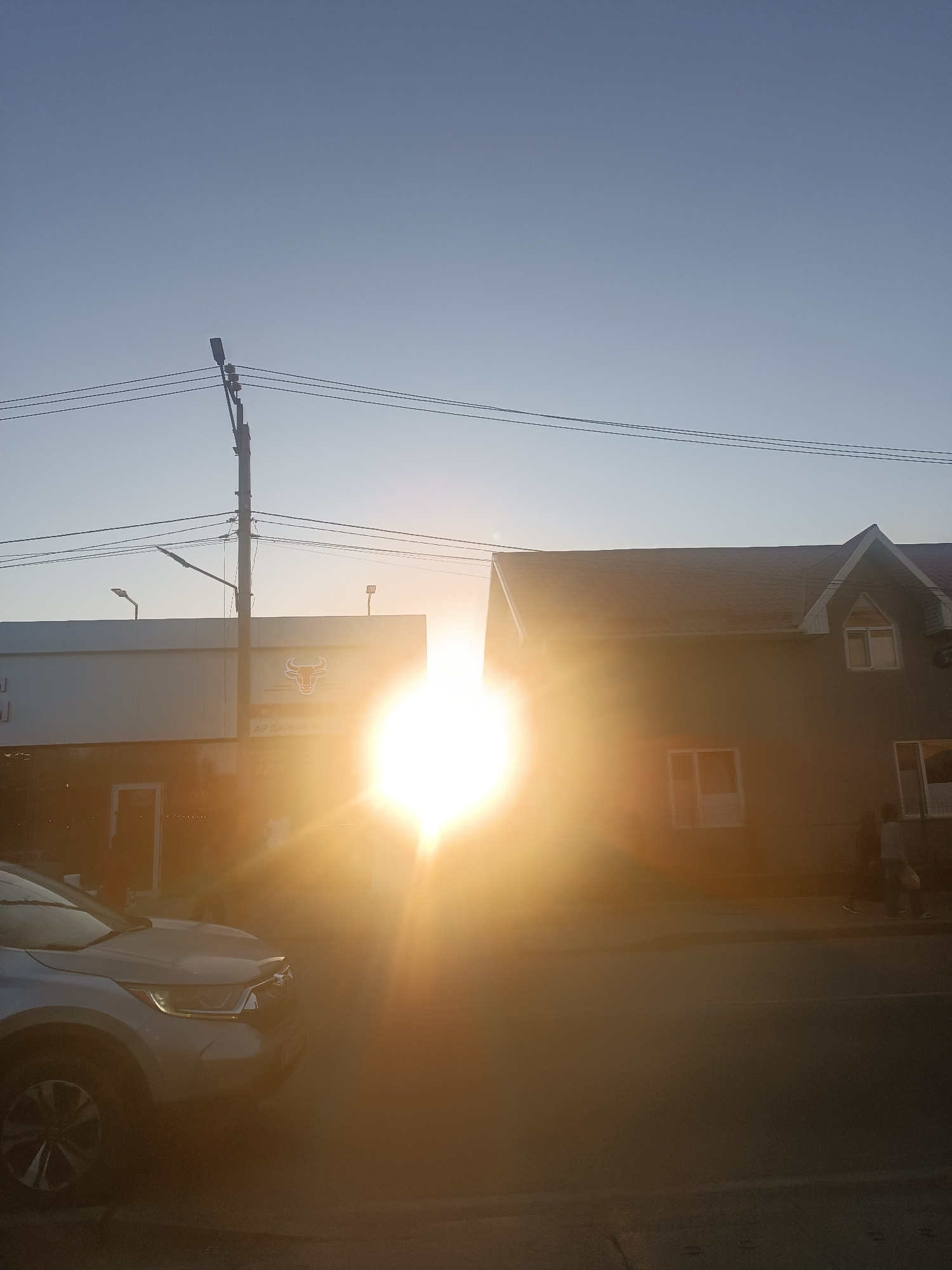 The image captures a street scene in Novopavlovka, Kyrgyzstan, during a bright sunset. The sun is positioned in the center of the frame, creating a strong lens flare that obscures details. To the left, a white building with a bull's head logo is visible, and to the right, a two-story building with dark siding and peaked roofs is present. Power lines stretch across the upper part of the image. A silver car is parked on the left side of the street, partially obscured by the sun's glare. In the distance, two individuals are walking on the sidewalk in front of the right building. The sky is clear and blue, transitioning to a lighter shade near the horizon.