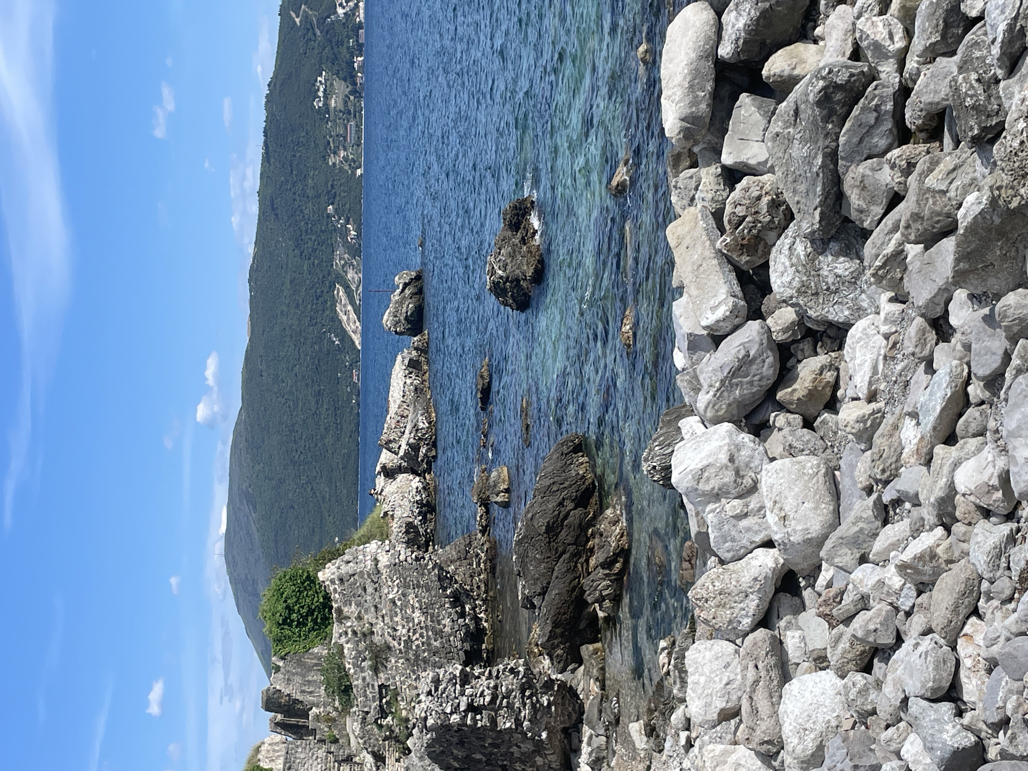 This image depicts a vibrant coastal landscape in Herceg Novi, Montenegro, presented under a bright, clear blue sky. The scene is static, capturing the natural beauty of the area during what appears to be late morning or early afternoon.
In the foreground, a rugged shoreline is visible, characterized by a dense accumulation of light-colored, predominantly grey and white, rough-hewn rocks and boulders of various sizes. This rocky embankment meets the clear, blue-green waters of the sea.
The middle ground showcases the transparent sea, where the light filtering through the surface reveals the rocky seabed and submerged rock formations. Several darker, craggy rocks emerge from the water, creating natural focal points. The surface of the water is gently rippled, suggesting calm conditions. Along the left edge of the frame, close to the waterline, lie the remnants of old stone structures, possibly ruins of a wall or building, partially overgrown with green shrubbery.
Further in the background, a large, undulating mountain range rises, densely covered in dark green foliage. On its slopes, particularly towards the right, small, light-colored buildings are sparsely scattered, indicating distant human settlements integrated into the natural environment. The sky above is a brilliant blue, punctuated by a few wispy white clouds.
No people are clearly visible in the image, and there is no discernable text. The overall impression is one of serene natural beauty combined with a sense of history provided by the ancient ruins.