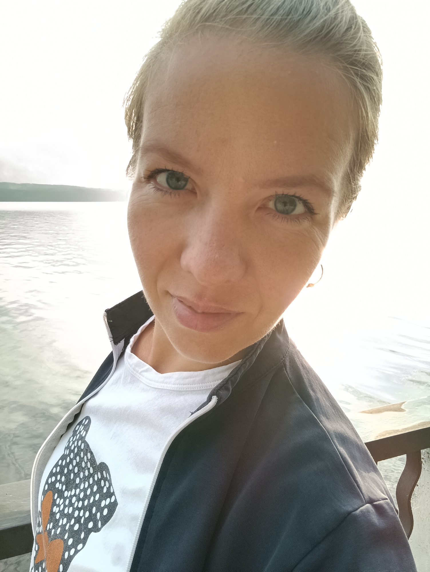 A young woman with short, blonde hair takes a selfie outdoors. She is wearing a white t-shirt with a black and white dotted pattern and an open dark blue jacket. Her head is tilted slightly to her left, and she is looking directly at the camera with a slight smile. She has light blue eyes and fair skin. Behind her, a body of water, likely a river or lake, reflects the bright sky. The water appears calm with some gentle ripples. In the distance, the hazy silhouette of a shoreline with trees is visible. The image is taken in daylight, with the sun positioned behind the woman, creating a slight flare effect. The background suggests a natural, possibly park-like or waterfront area in Chaikovsky, Russia.