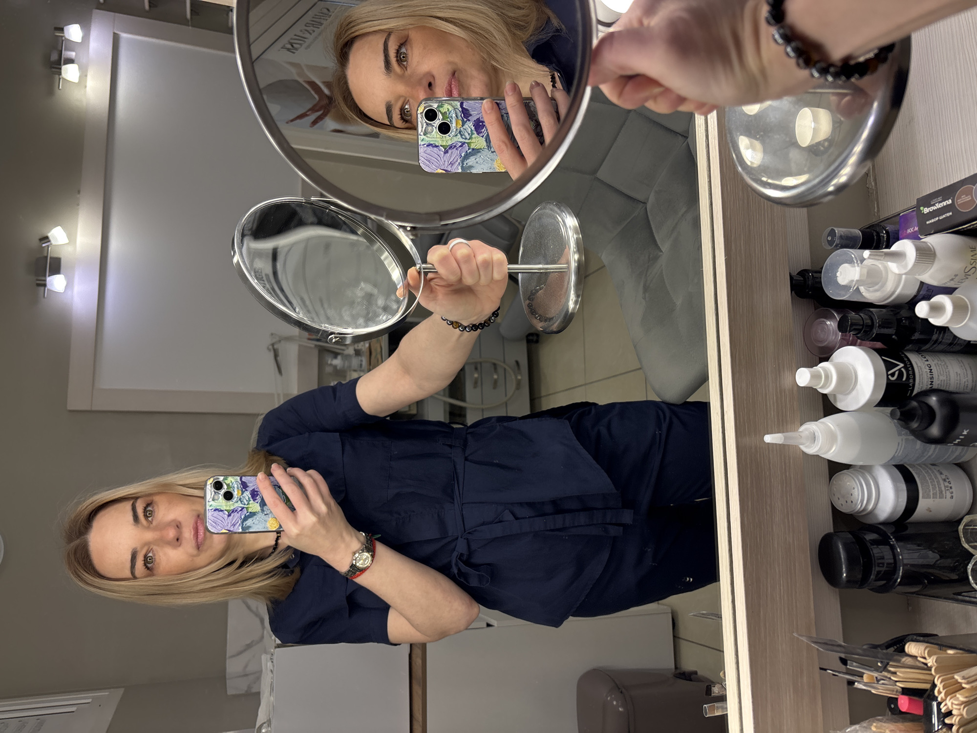 A woman is taking a selfie in a mirror. She is holding a phone in her right hand with a case that has a blue and purple floral design. She is wearing a dark blue outfit with a tie around the waist and a watch on her left wrist with a red band. Her hair is blonde and she has light eyes. In the background, there is a wall with two light fixtures on it. There is also another mirror being held by her other hand. On a counter to the right of the frame, there are various bottles and containers, along with wooden sticks and a dark box. The scene is illuminated by artificial lighting. The location appears to be indoors, possibly a salon or treatment room.
