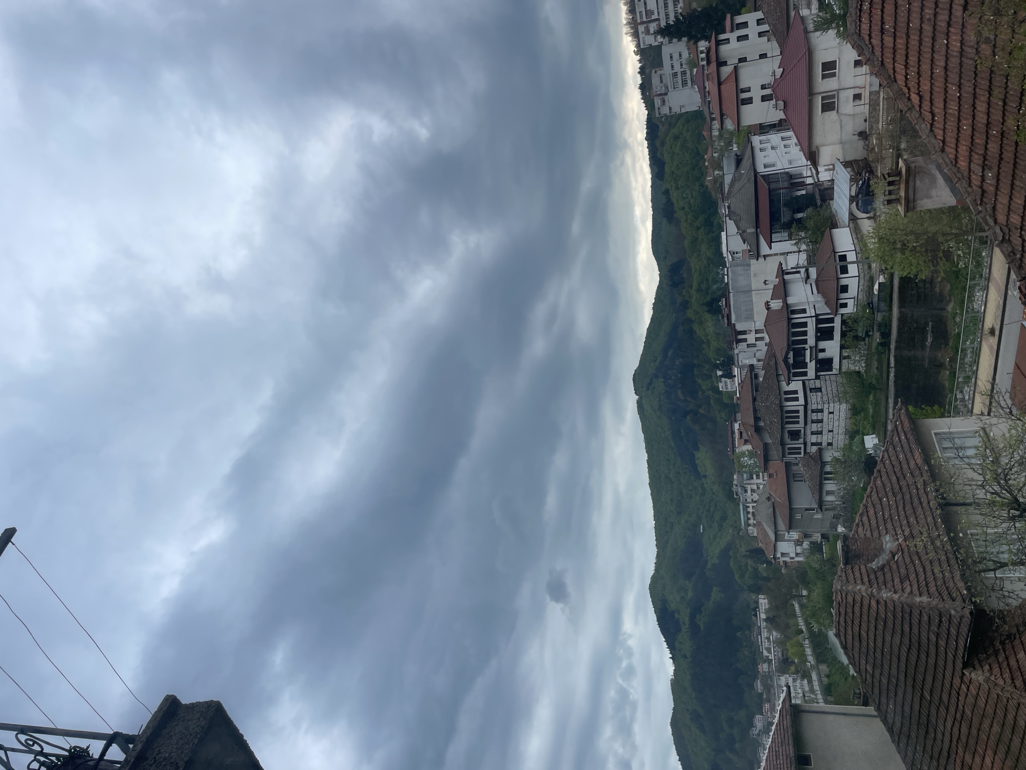 The image displays a panoramic view of a town nestled in a mountainous landscape under an overcast sky. The scene is captured from an elevated perspective, looking down and across the valley.
The upper half of the image is dominated by a dense, grey, and cloudy sky, indicating heavily overcast weather. There are variations in cloud thickness, with lighter patches suggesting diffused sunlight attempting to break through, particularly along the horizon line above the mountains.
A prominent, verdant mountain range runs horizontally across the mid-ground, completely covered in dense green trees, suggesting a lush forest. The town's buildings are situated along the lower slopes and at the base of this mountain, stretching across the middle and lower parts of the frame. The architecture features numerous multi-story houses with predominantly white or light-colored facades and distinct reddish-brown tiled roofs, typical of the region. The houses appear closely packed, reflecting the characteristics of a town like Zlatograd, Bulgaria.
In the immediate foreground, on the right side, a dark, reddish-brown tiled roof is visible, partially obscured, indicating the elevated position from which the photo was taken. On the far left foreground, a dark metal railing or structure is partially visible.
The scene appears static; there are no people or explicit activities visible. The time of day is clearly daytime, characterized by soft, diffused lighting due to the thick cloud cover. There is no visible text present within the image.