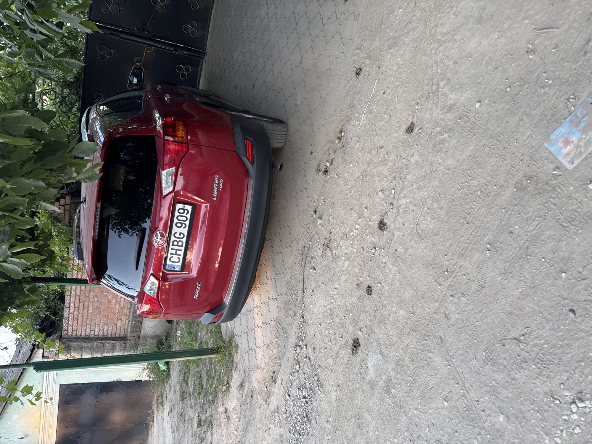 A red Toyota RAV4, bearing the license plate CHBG 909, is parked on a textured concrete and gravel surface. The vehicle is viewed from the rear, with its right side closer to a black metal gate. Adjacent to the car, a brick wall and a light green building with a dark garage door are visible. Lush green foliage hangs overhead, partially obscuring the view. The ground is uneven, with scattered pebbles and dark, circular debris that may be animal droppings. The scene appears to be an outdoor area, possibly a driveway or yard, in the city of Cahul, Moldova.