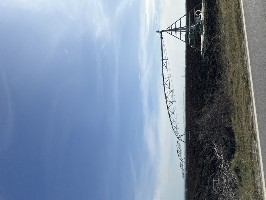 The image depicts an expansive agricultural landscape in Karanovo, Bulgaria, under a clear blue sky with sparse, wispy white clouds. A multi-section center pivot irrigation system dominates the midground, extending horizontally across a vast, dark brown, tilled field. The irrigation system's pivot point, a triangular metal tower supporting machinery and a base platform with a white coiled hose, is located on the right. Multiple truss-like metal sections, each supported by wheels, arc from this pivot towards the left, with further segments receding into the distant horizon. In the immediate foreground, a strip of asphalt road runs across the bottom of the frame, bordered by a dry, grassy verge where bare branches and dark, thorny bushes are present on the left. No individuals or active processes are visible in the scene.